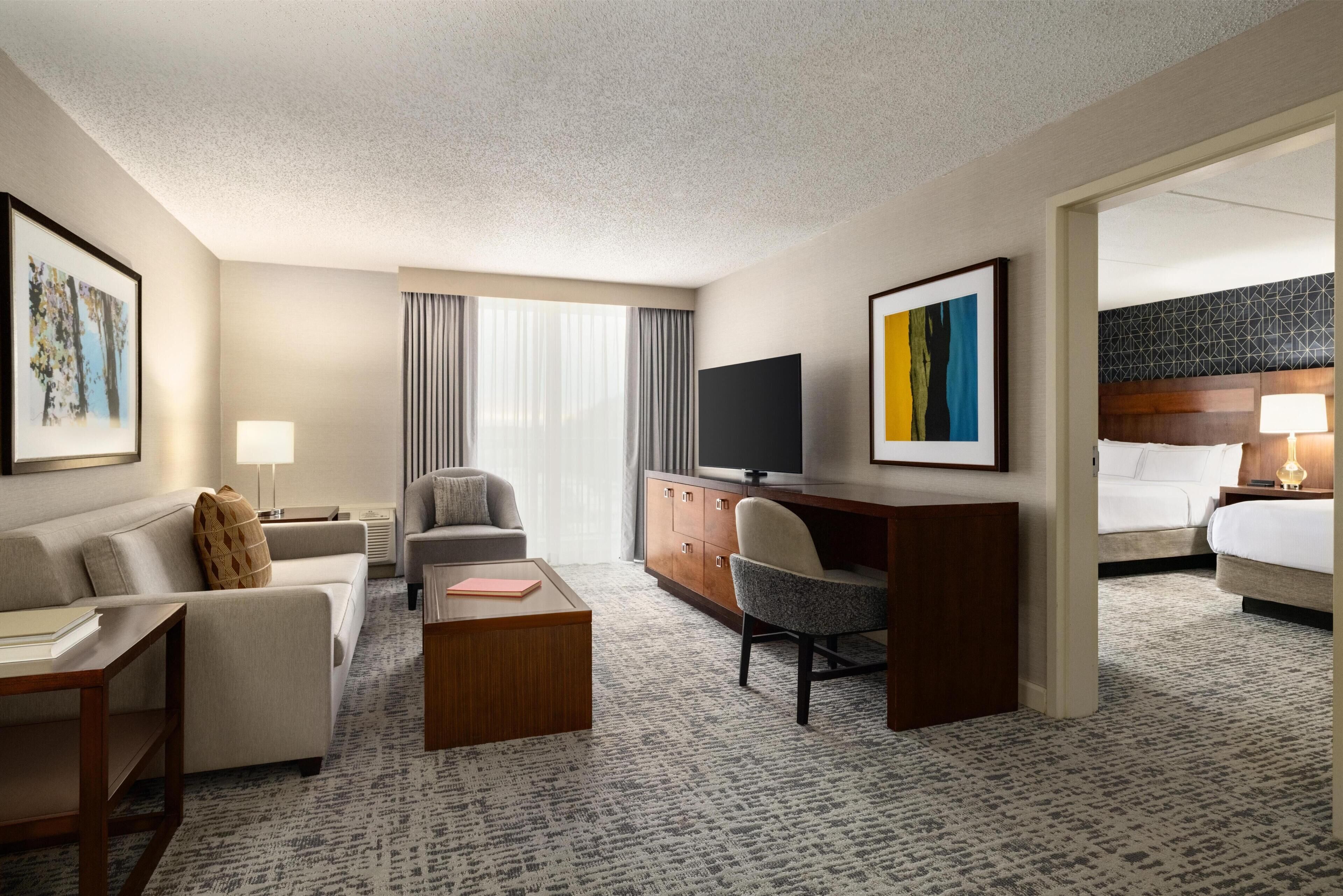 A hotel room with a sofa, armchair, desk, television, and a bed.