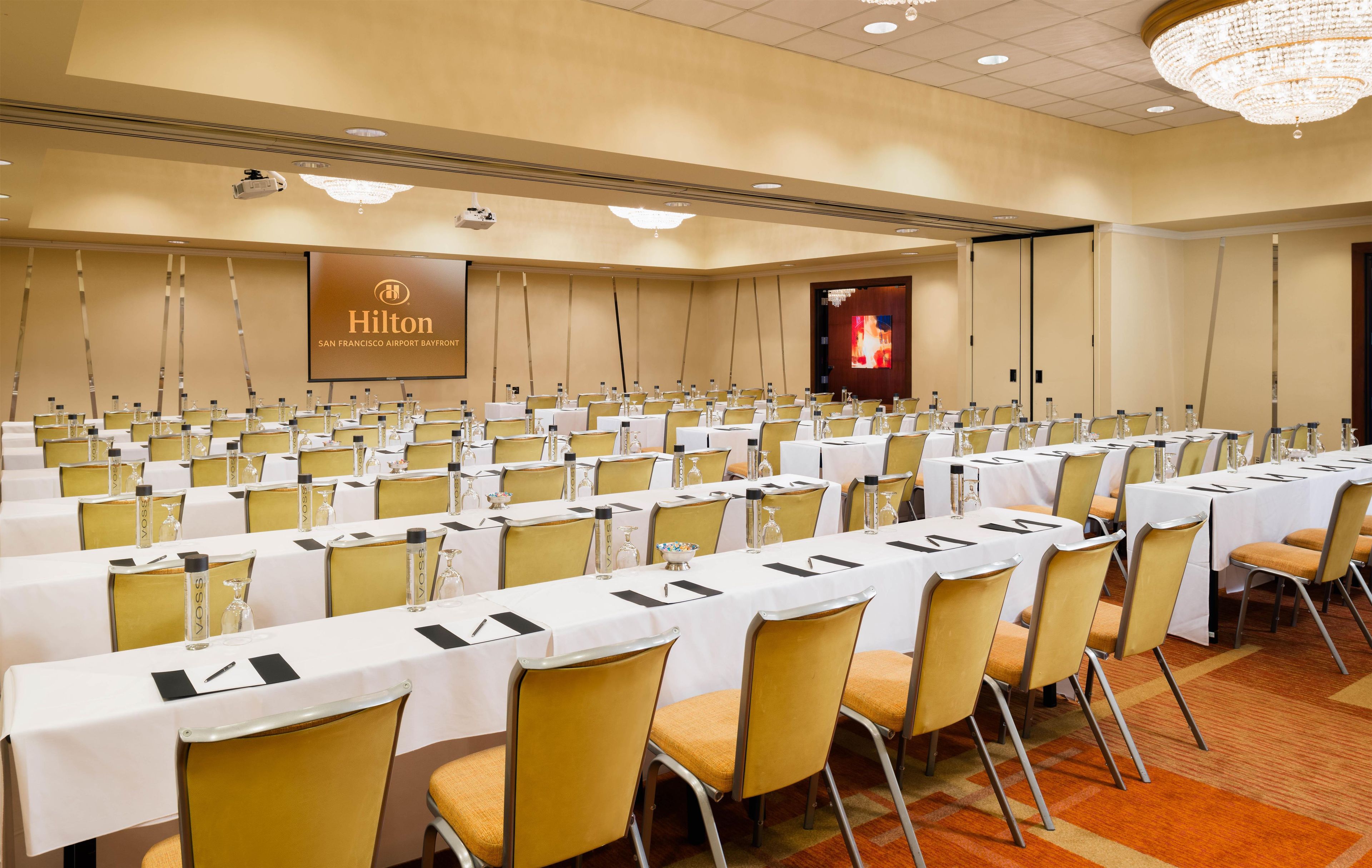 A conference room with rows of tables, chairs, and a large screen displaying the Hilton logo.