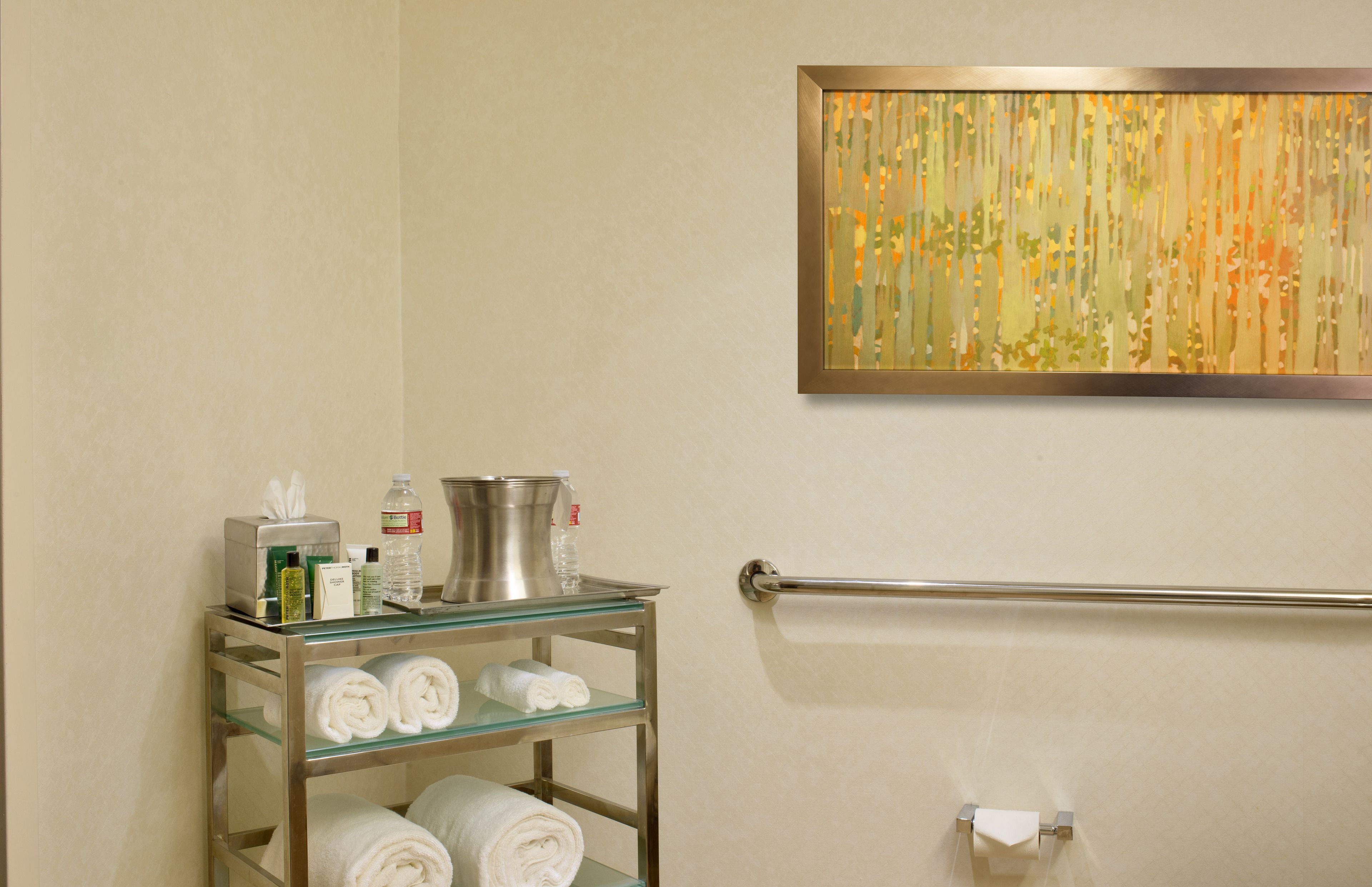 A bathroom with a glass shelf holding towels, toiletries, and a metal pitcher.