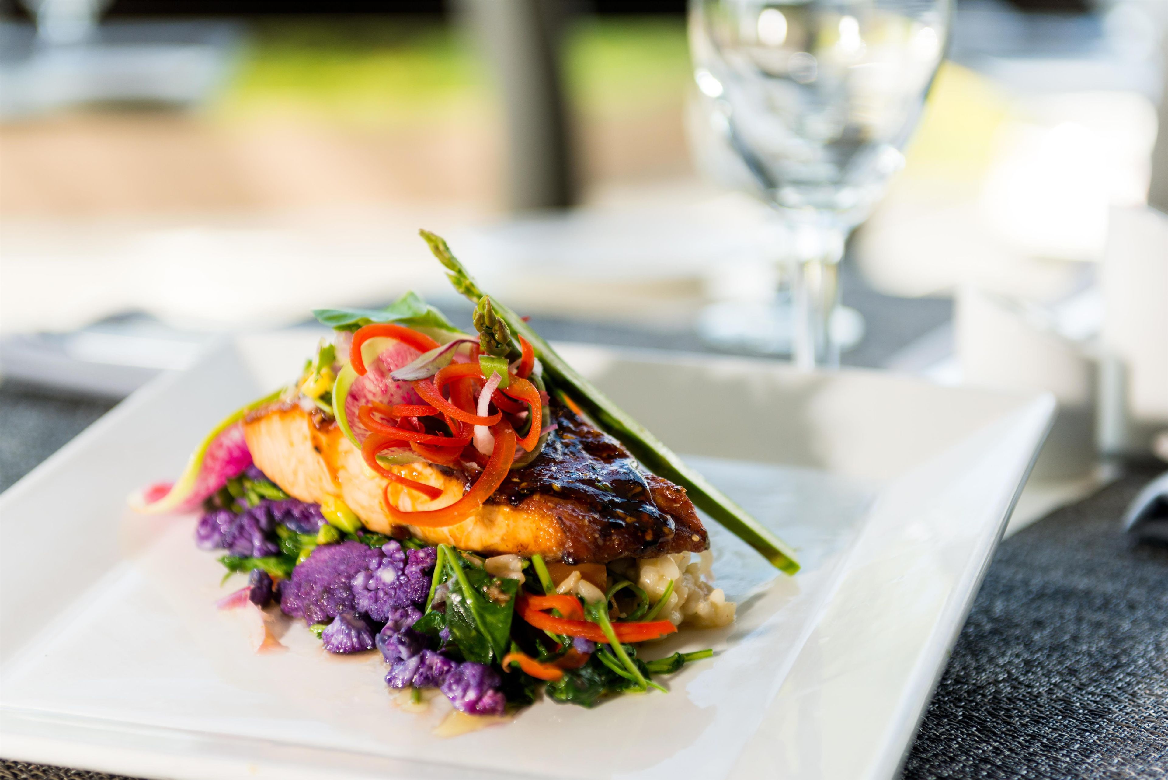 A plated dish with a variety of vegetables and a piece of meat, possibly salmon, on a rectangular white plate.