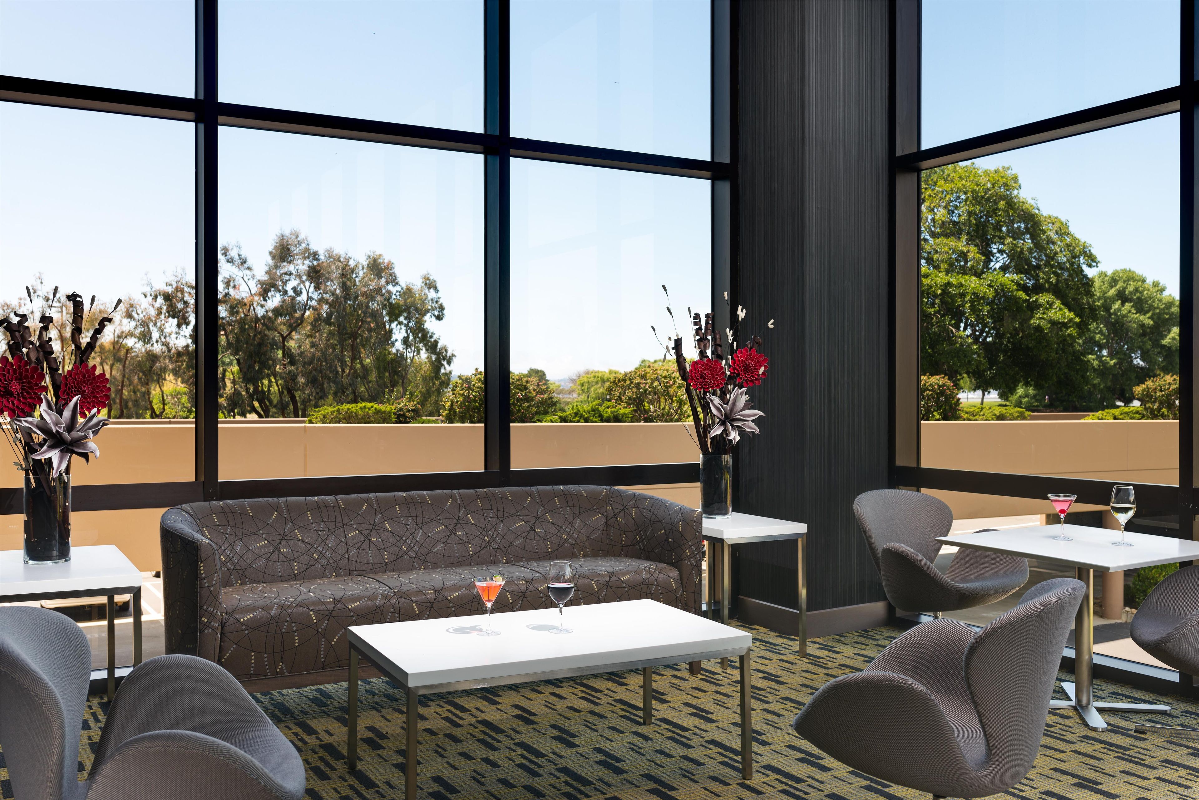 A modern lounge area with a sofa, chairs, and a table, featuring large windows and a view of greenery.