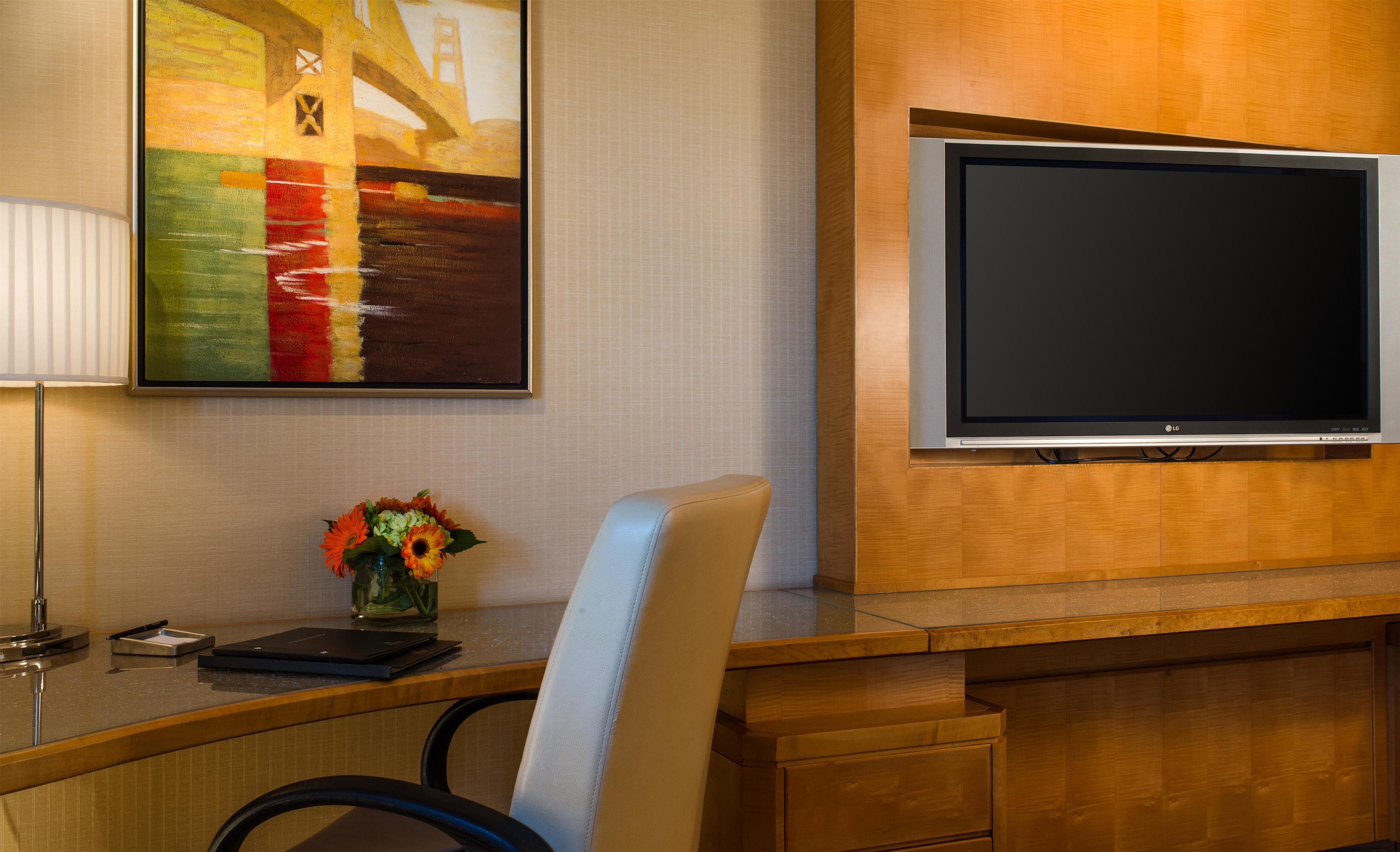 Habitación de hotel con televisor de pantalla plana en la pared, un escritorio con una computadora portátil, un jarrón con flores y un cuadro sobre el escritorio.