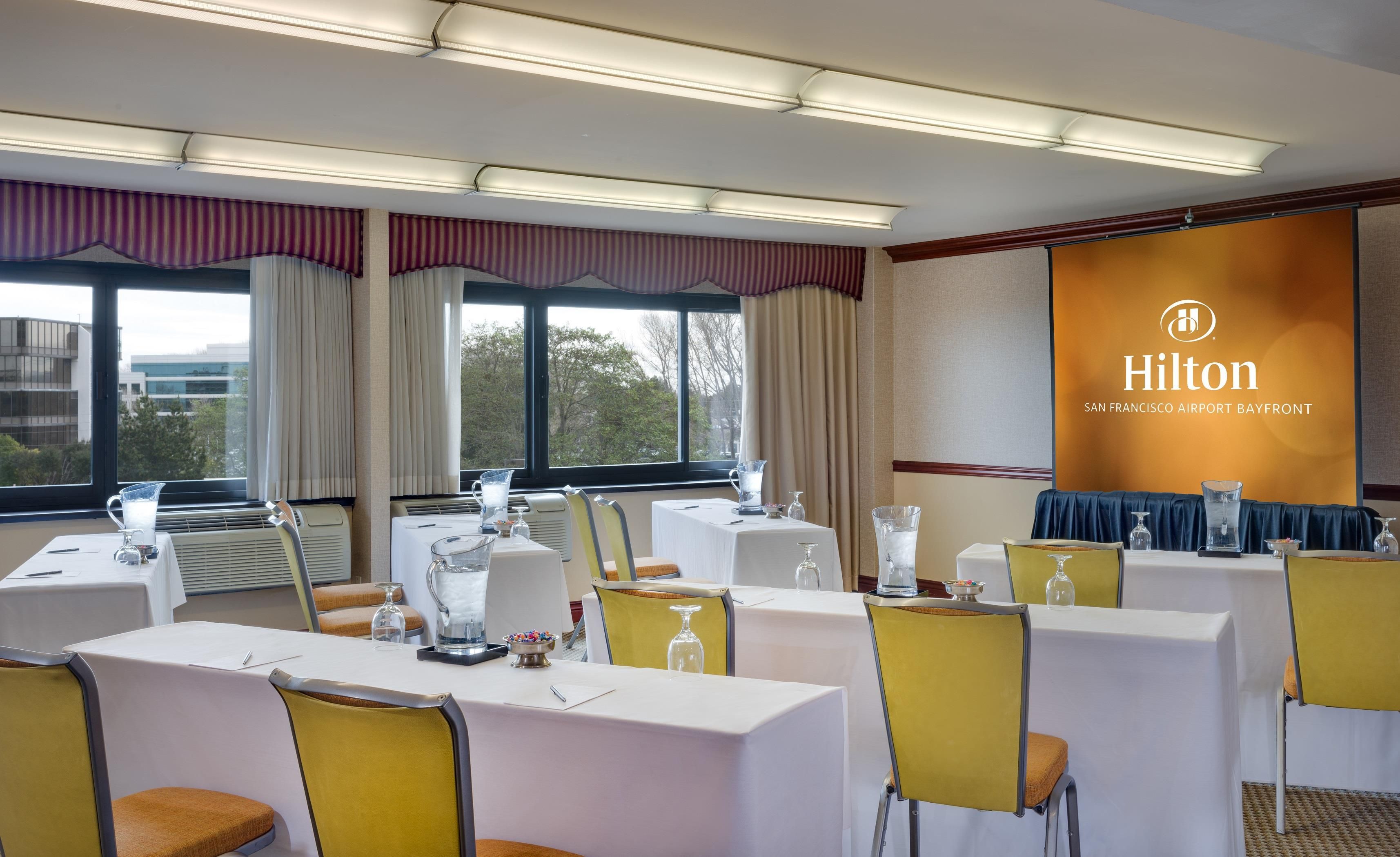 Sala de conferências com mesas, cadeiras e tela de projeção no Hilton San Francisco Airport Bayfront.