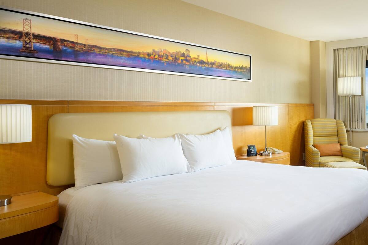 A hotel room with a large bed, a headboard, a nightstand, a chair, and a painting on the wall.