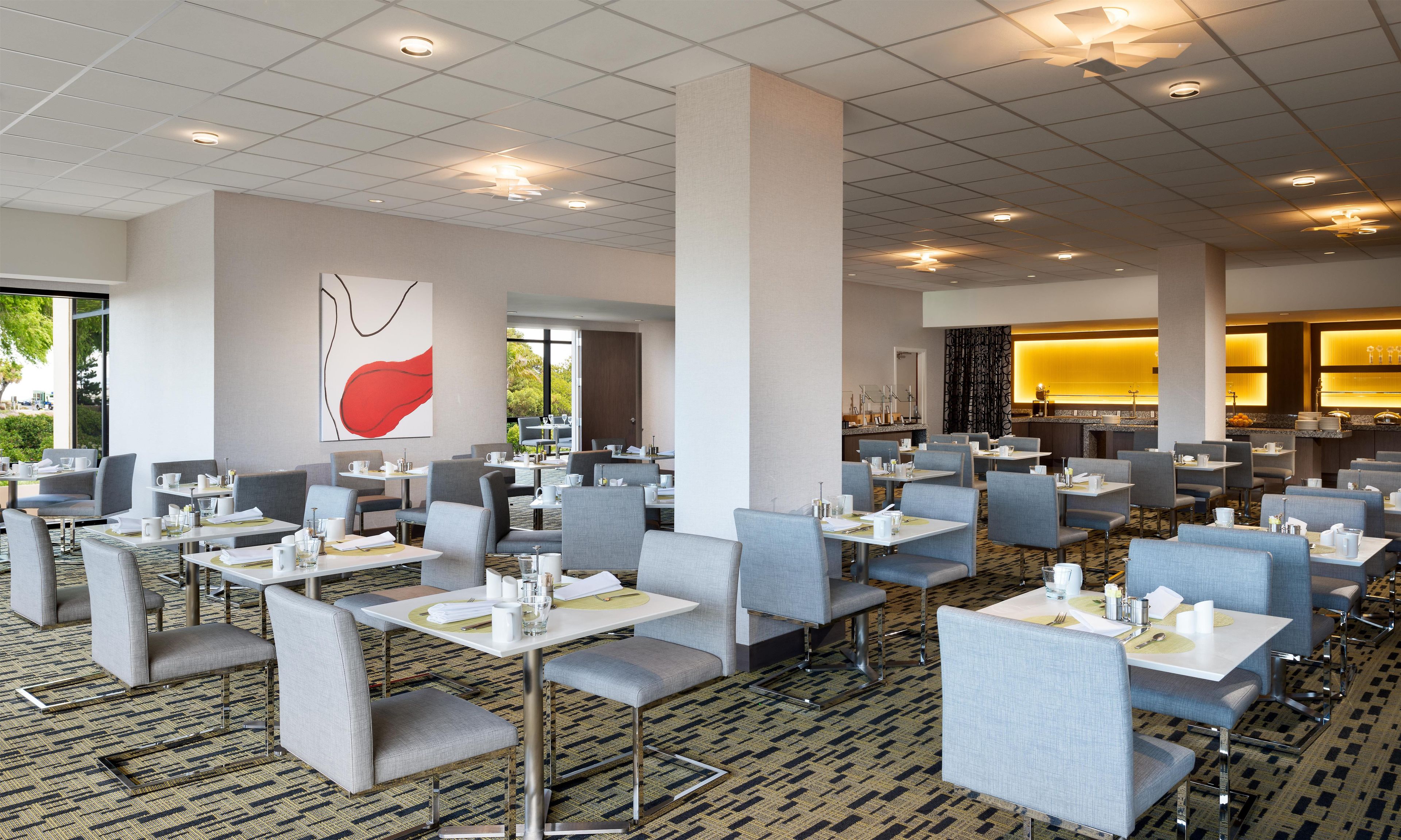Une salle à manger d’hôtel avec de nombreuses tables et chaises, une grande peinture abstraite au mur et une vue sur l’extérieur grâce à de grandes fenêtres.