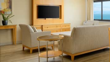 A modern hotel room with a flat-screen TV mounted on a wooden panel, a beige sofa, a round glass coffee table, and a view of the sea through a large window.