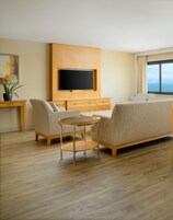 Una habitación de hotel moderna con un televisor de pantalla plana montado en un panel de madera, un sofá color beige, una mesa de centro redonda de vidrio y vistas al mar a través de un ventanal grande.