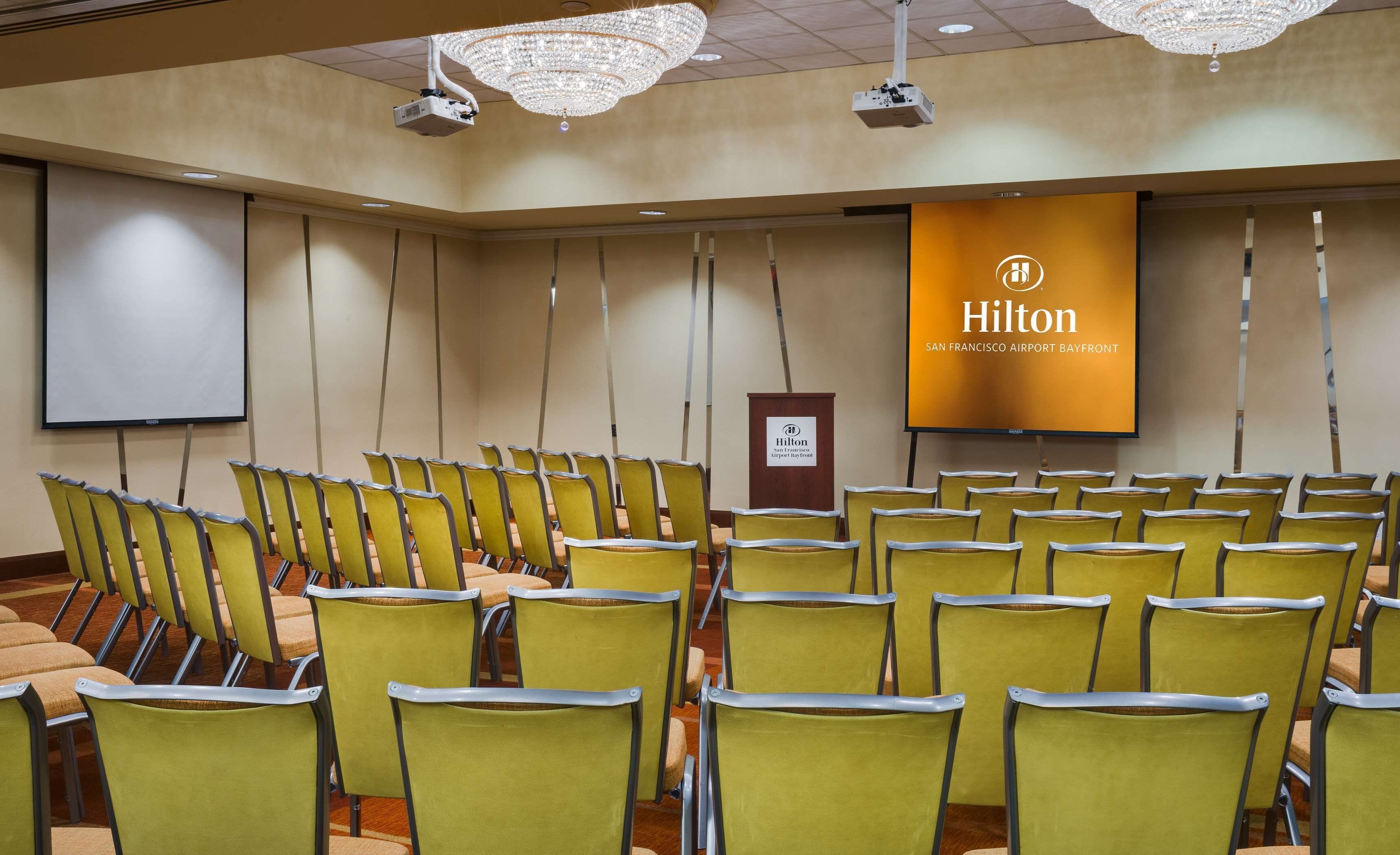 A conference room with rows of yellow chairs, a large screen, and a Hilton banner.