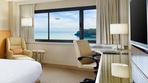 Et hotelværelse med et stort vindue, der giver udsigt over havet og et fly i det fjerne. Sengen, to stole, et skrivebord med lampe og et fjernsyn.