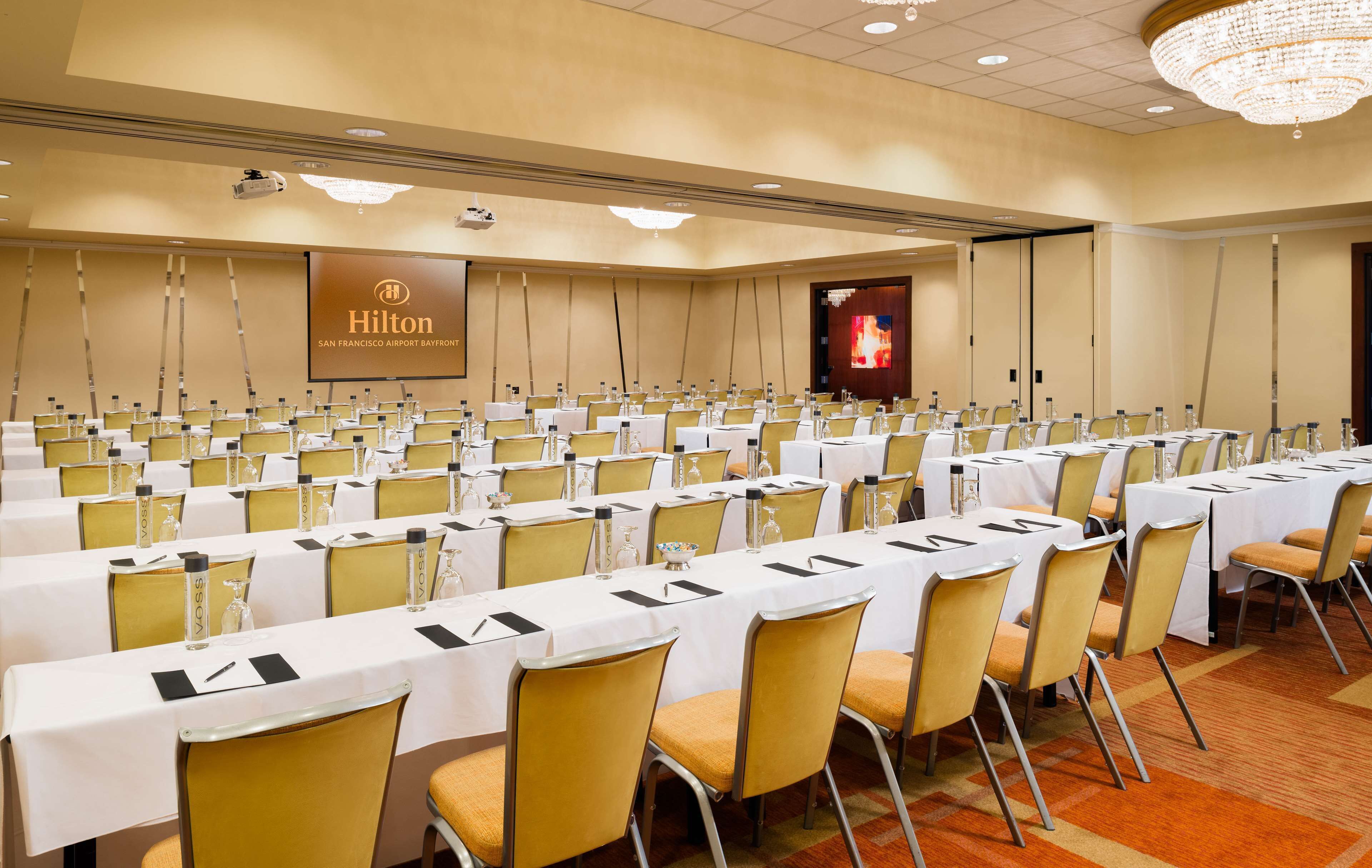 A conference room with rows of tables and chairs, a large screen, and a chandelier.