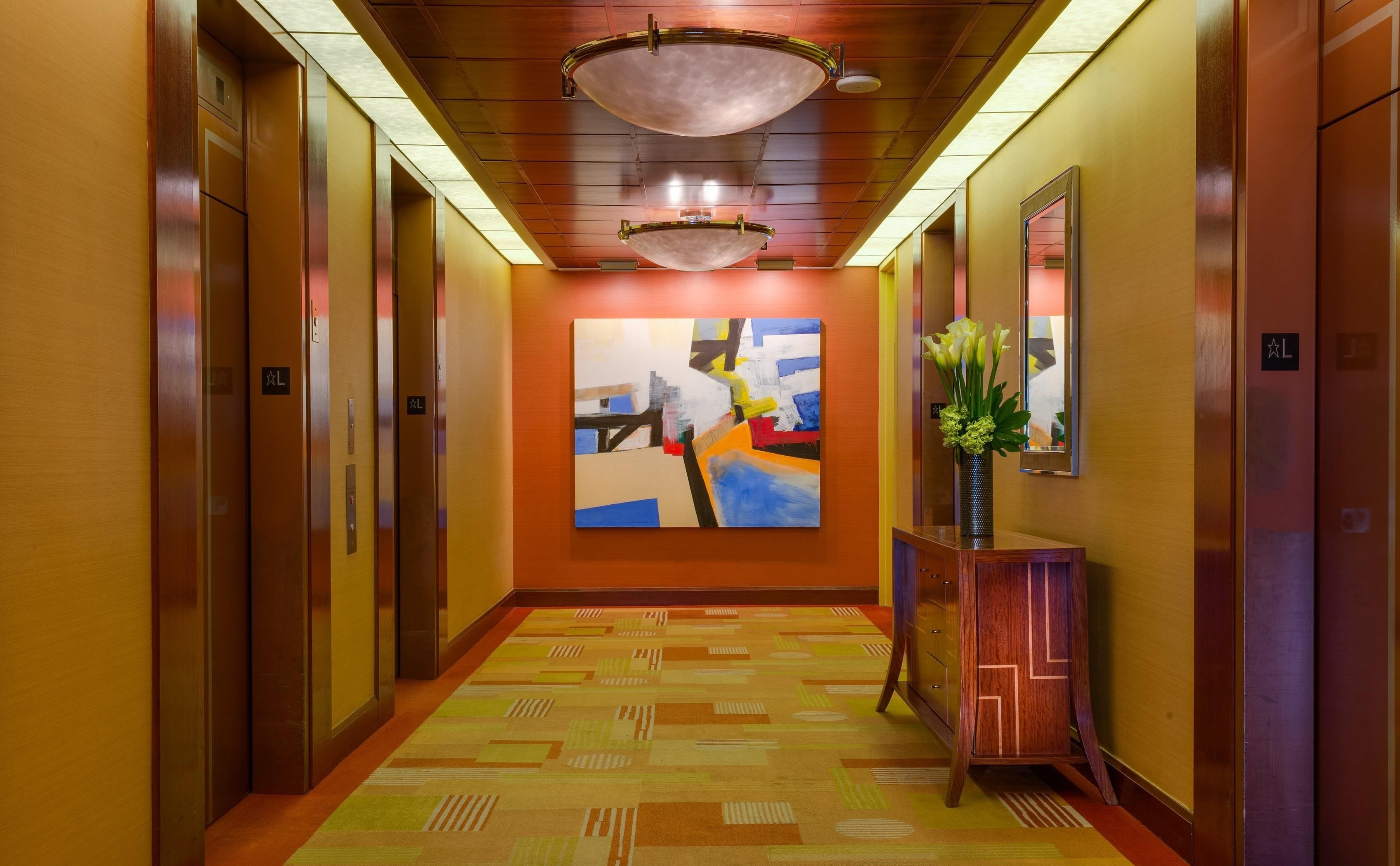 Un pasillo de hotel con ascensores, una alfombra estampada, un cuadro en la pared y un jarrón con flores.