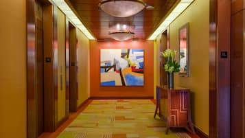Un pasillo de hotel con ascensores, una alfombra estampada, un cuadro en la pared y un jarrón con flores.
