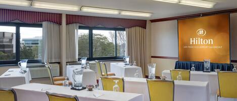 A conference room with tables and chairs, a large window with curtains, and a wall with the Hilton logo.