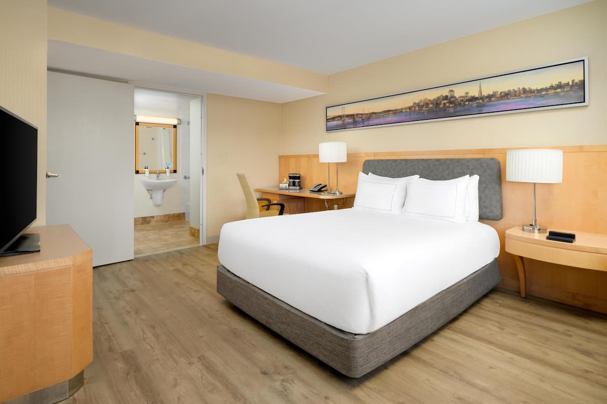 A hotel room with a large bed, bedside tables, a desk, a chair, a television, and a bathroom visible through an open door.