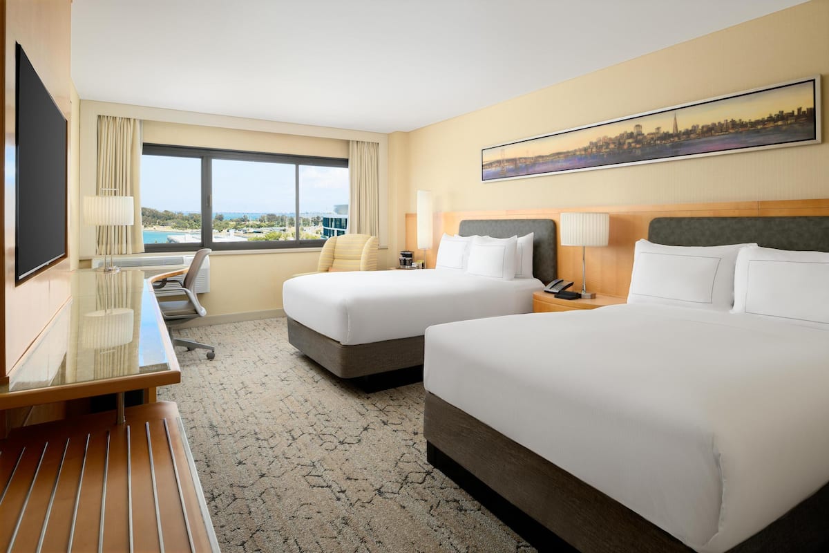 A hotel room with two beds, a desk, a chair, a TV, and a large window with a cityscape view.