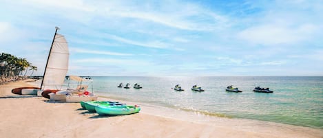 A beach with a sailboat, several jet skis, and a row of palm trees.