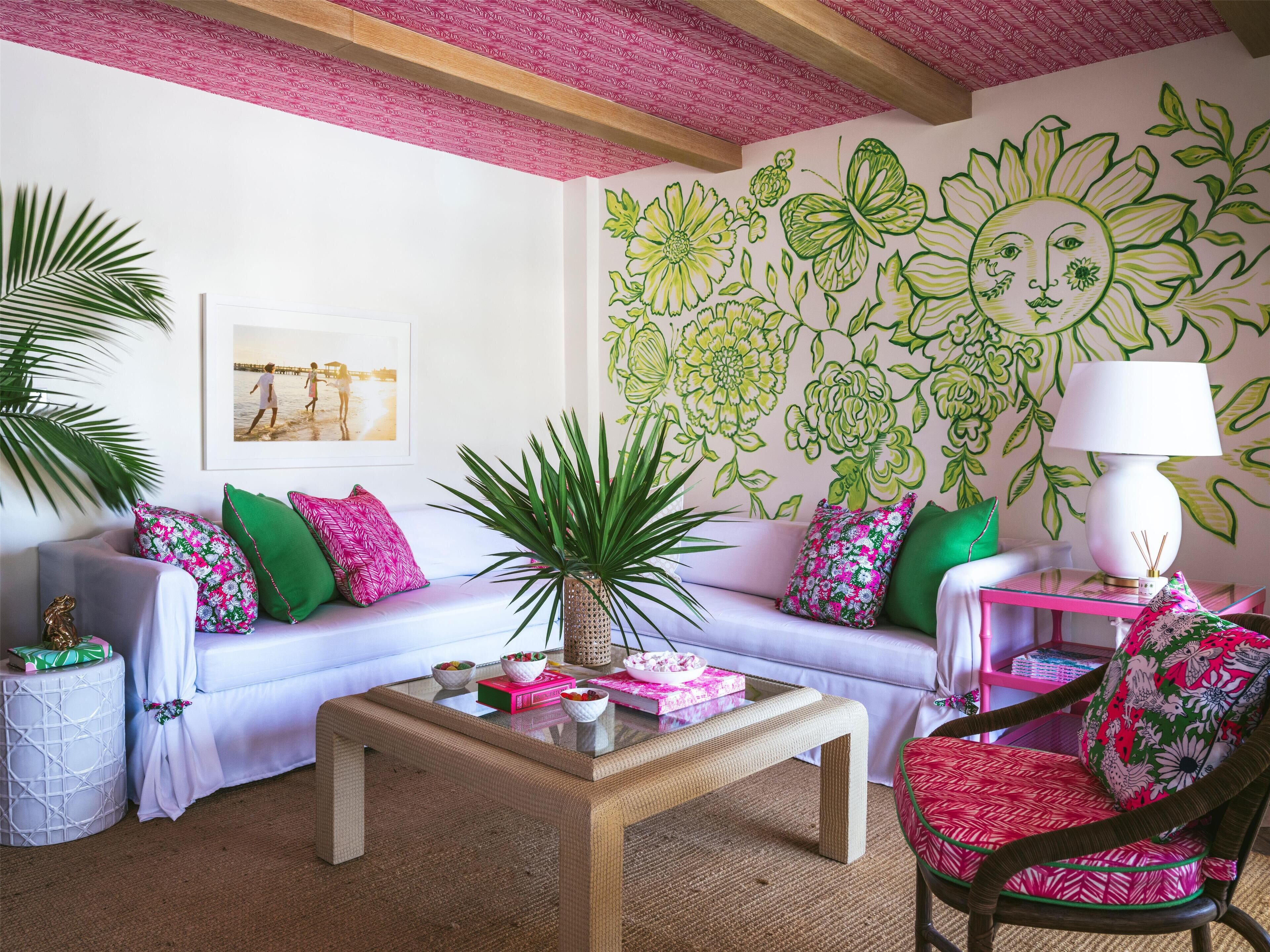 A cozy living room with a white sofa, green and pink cushions, a glass coffee table, and a chair with a red and white patterned cushion. A large floral wall mural and a framed picture are visible.