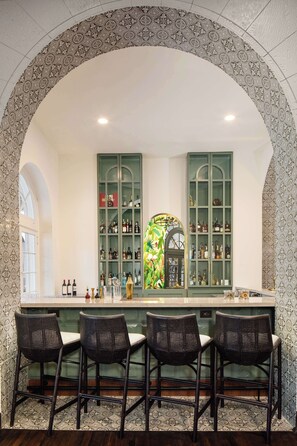 A bar area with a high, arched ceiling and patterned wallpaper. There are three wicker bar stools and a green cabinet with shelves of bottles.