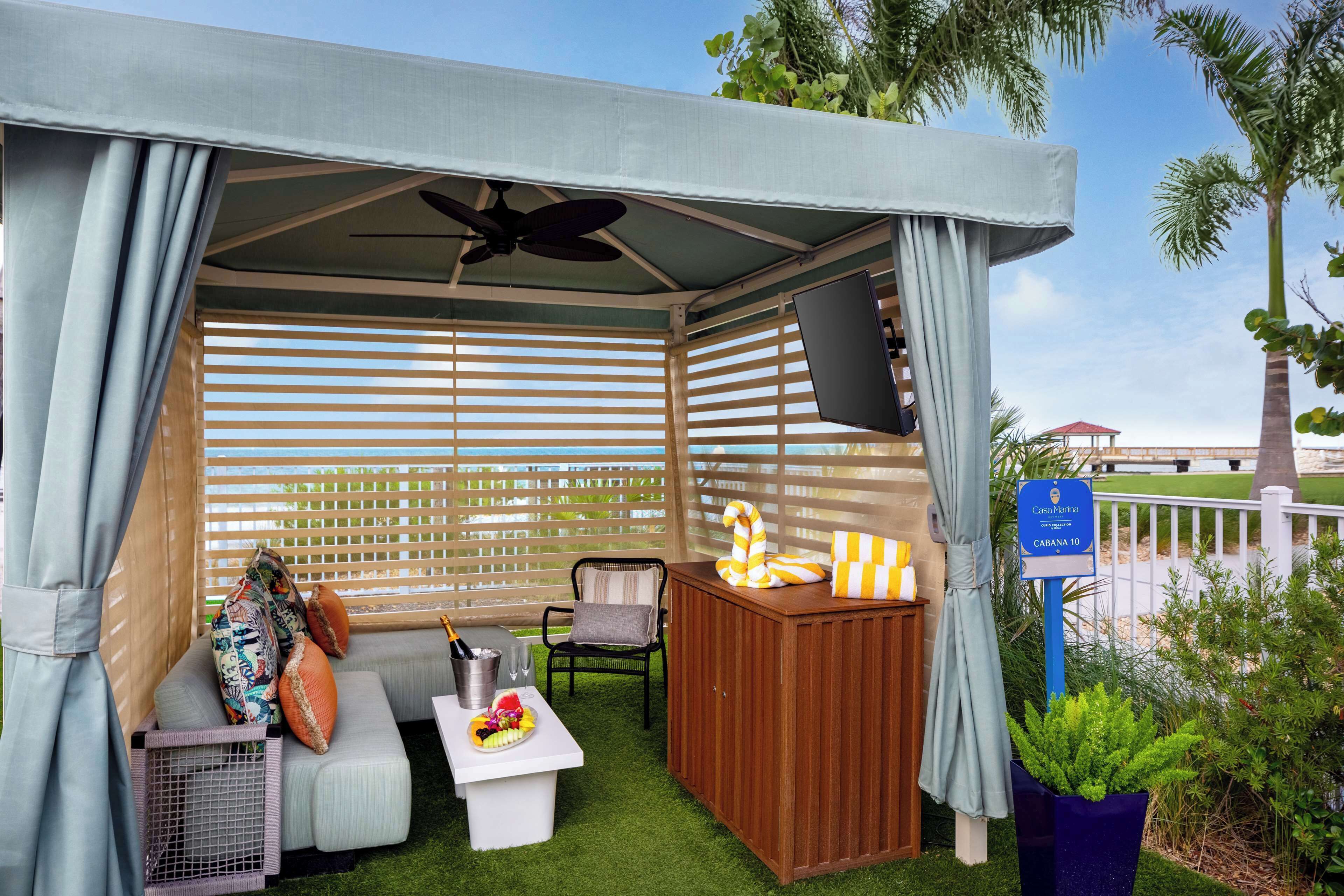 A covered outdoor seating area with a ceiling fan, a wooden table with a fruit bowl, and a sofa with cushions.