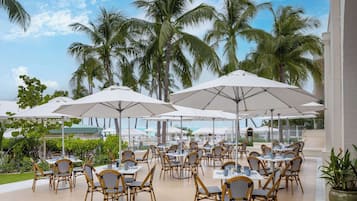 An outdoor dining area with tables and chairs under umbrellas, surrounded by palm trees.
