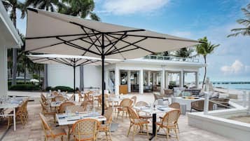 Un espace de restauration en plein air avec des chaises et des tables en osier, de grands parasols et une vue sur l’océan.