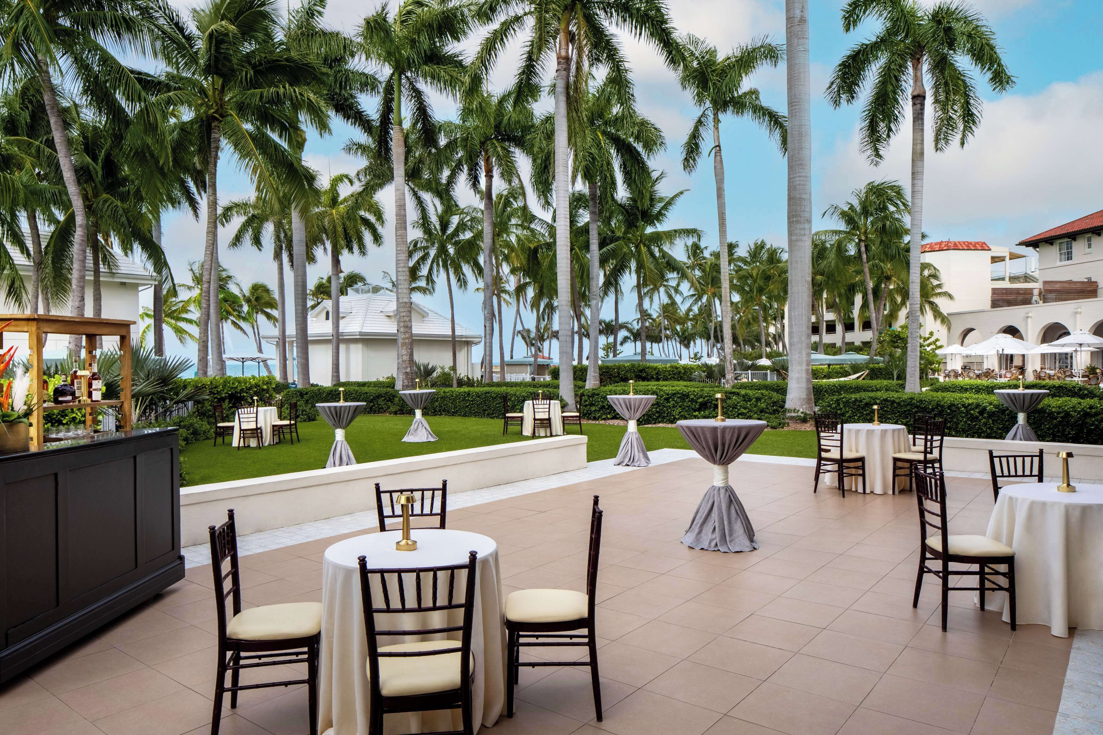 An outdoor dining area with round tables and chairs, surrounded by palm trees and a well-maintained lawn.