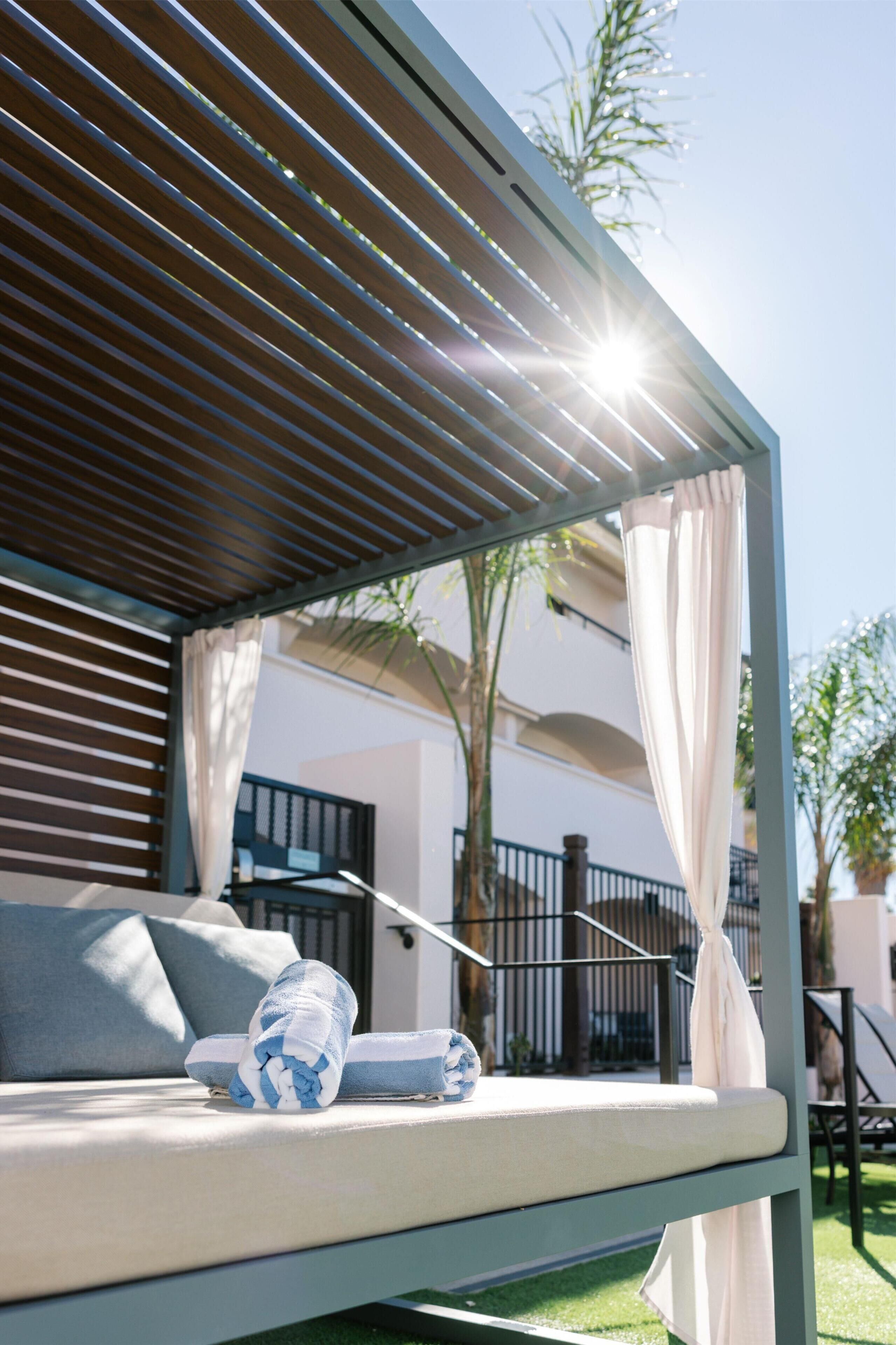 A sunlit outdoor seating area with a cushioned bench, a rolled towel, and a pair of slippers.