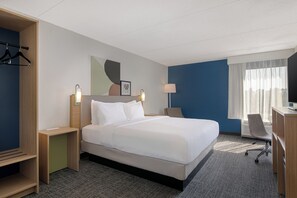 Una habitación de hotel con cama, escritorio, silla, armario y ventana con cortinas.