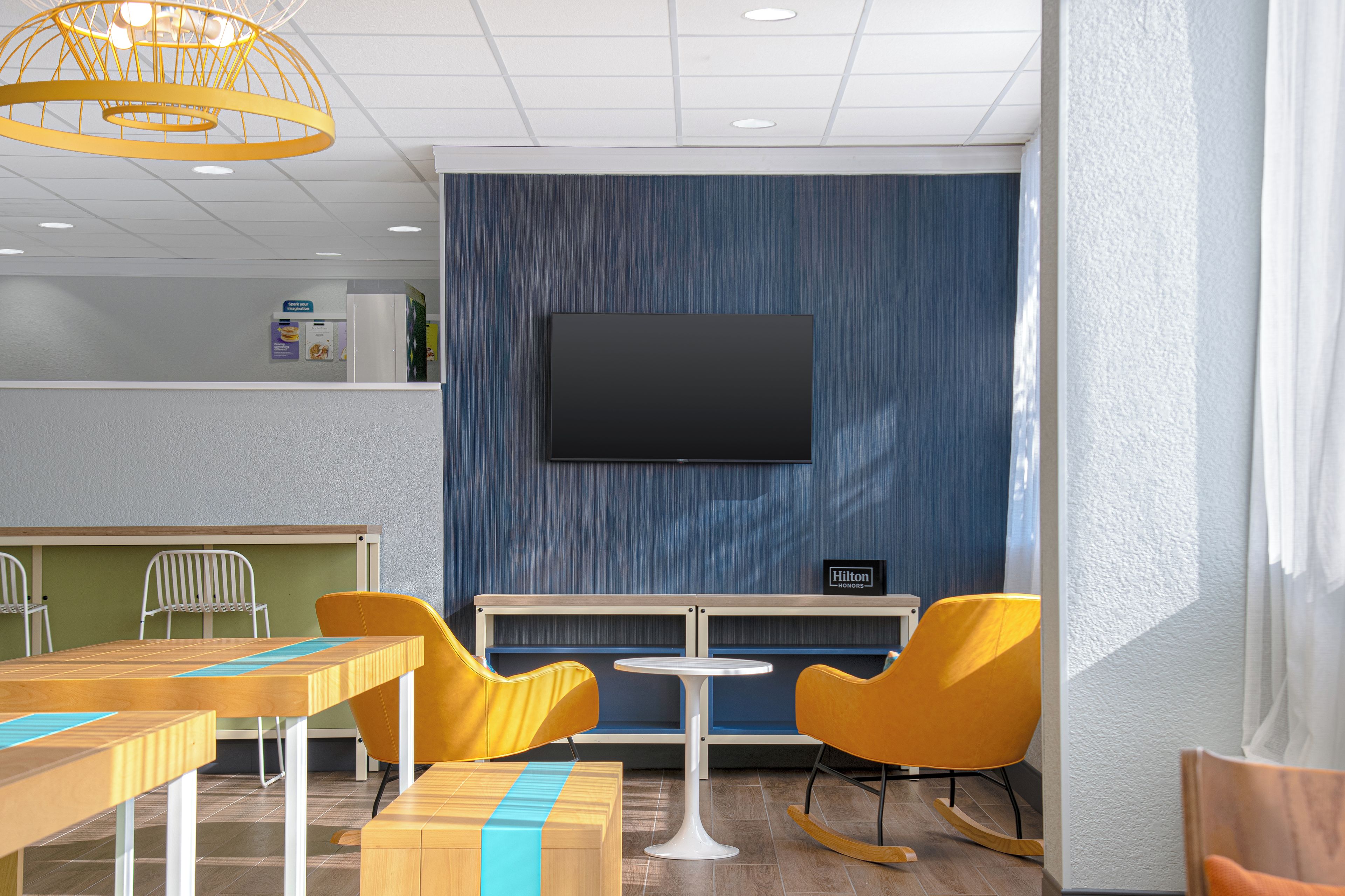 A hotel room with a flat-screen TV mounted on the wall, a small table with a chair, and a seating area with yellow chairs and a rocking chair.