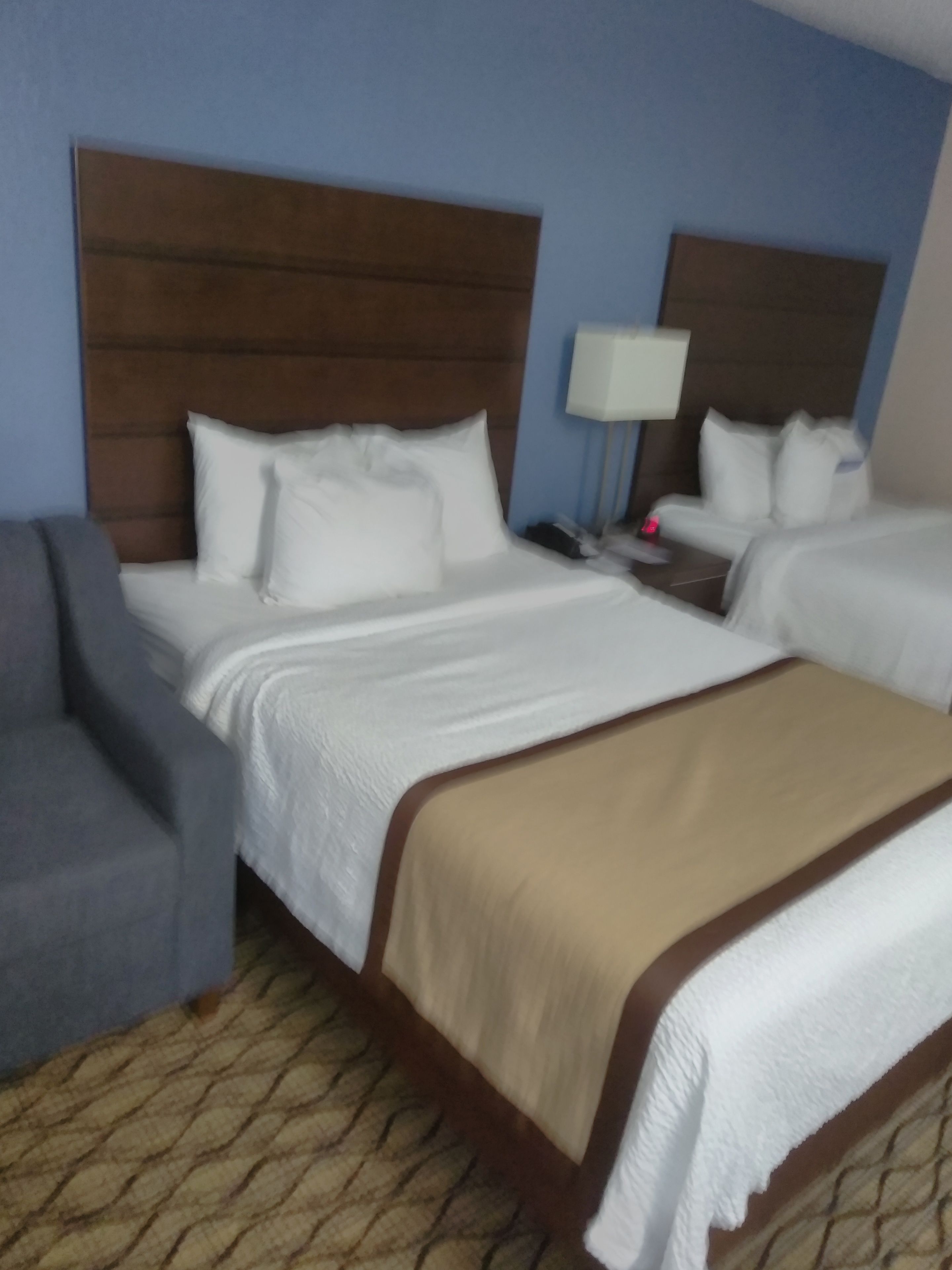 A hotel room with two beds, a nightstand with a lamp, and a gray armchair.