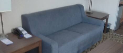 Una habitación con un sofá azul, una mesa de centro de madera, dos mesitas de noche con lámparas, un cuadro enmarcado en la pared y alfombra estampada.