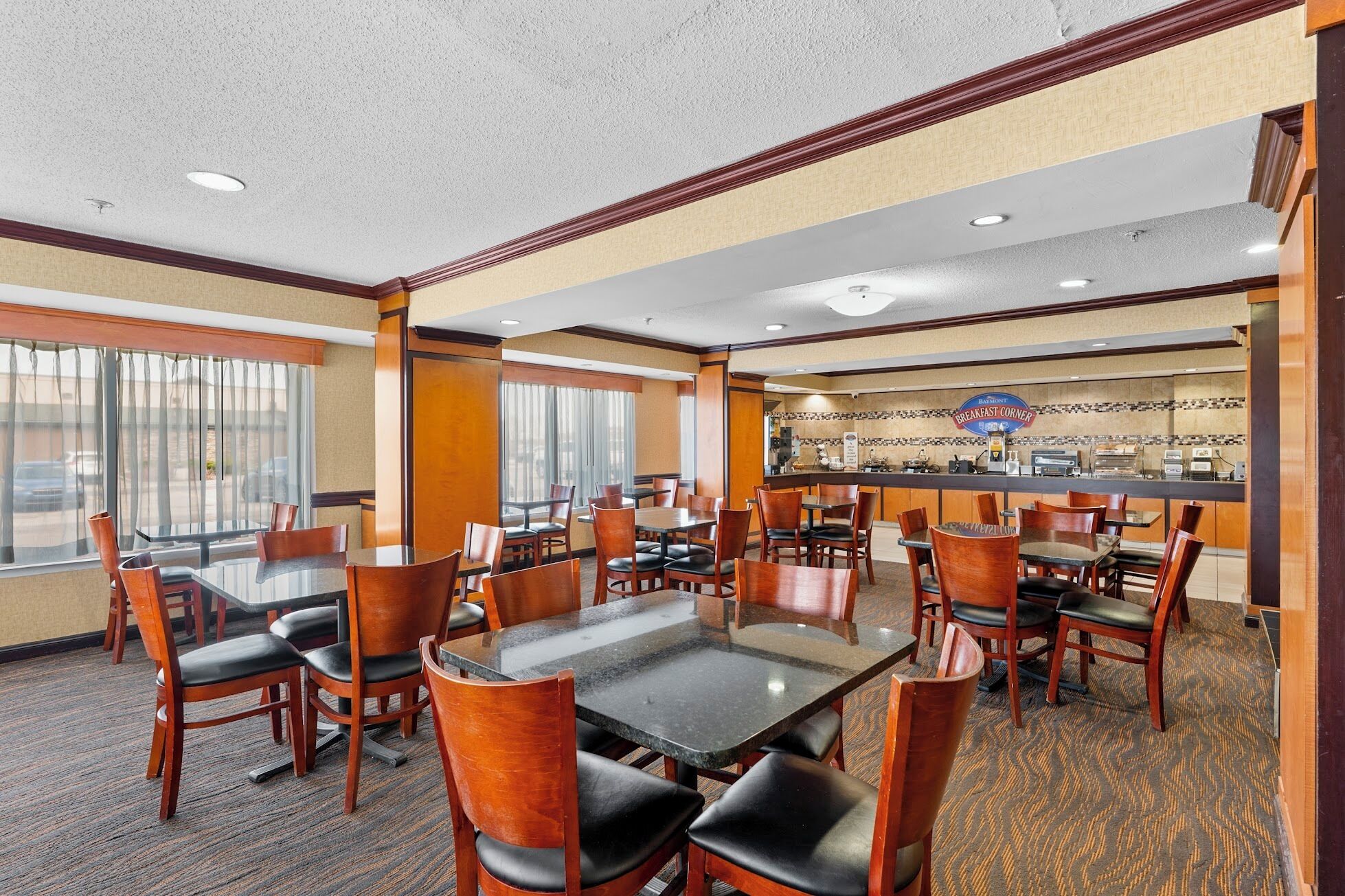 A hotel dining area with multiple tables and chairs, a breakfast counter, and large windows.