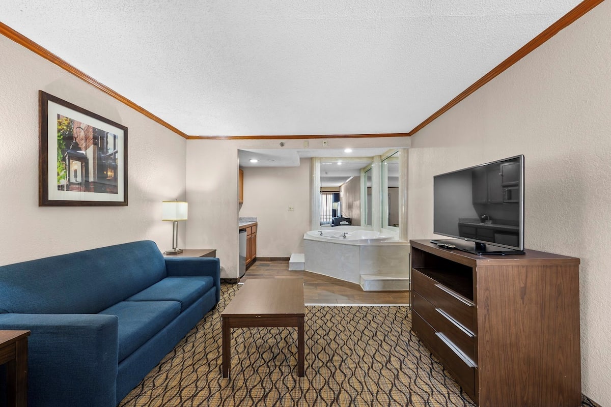 Ein Hotelzimmer mit einer Couch, einem Fernseher, einem kleinen Tisch, einem Spiegel und einem durch eine offene Tür einsehbaren Badezimmer.