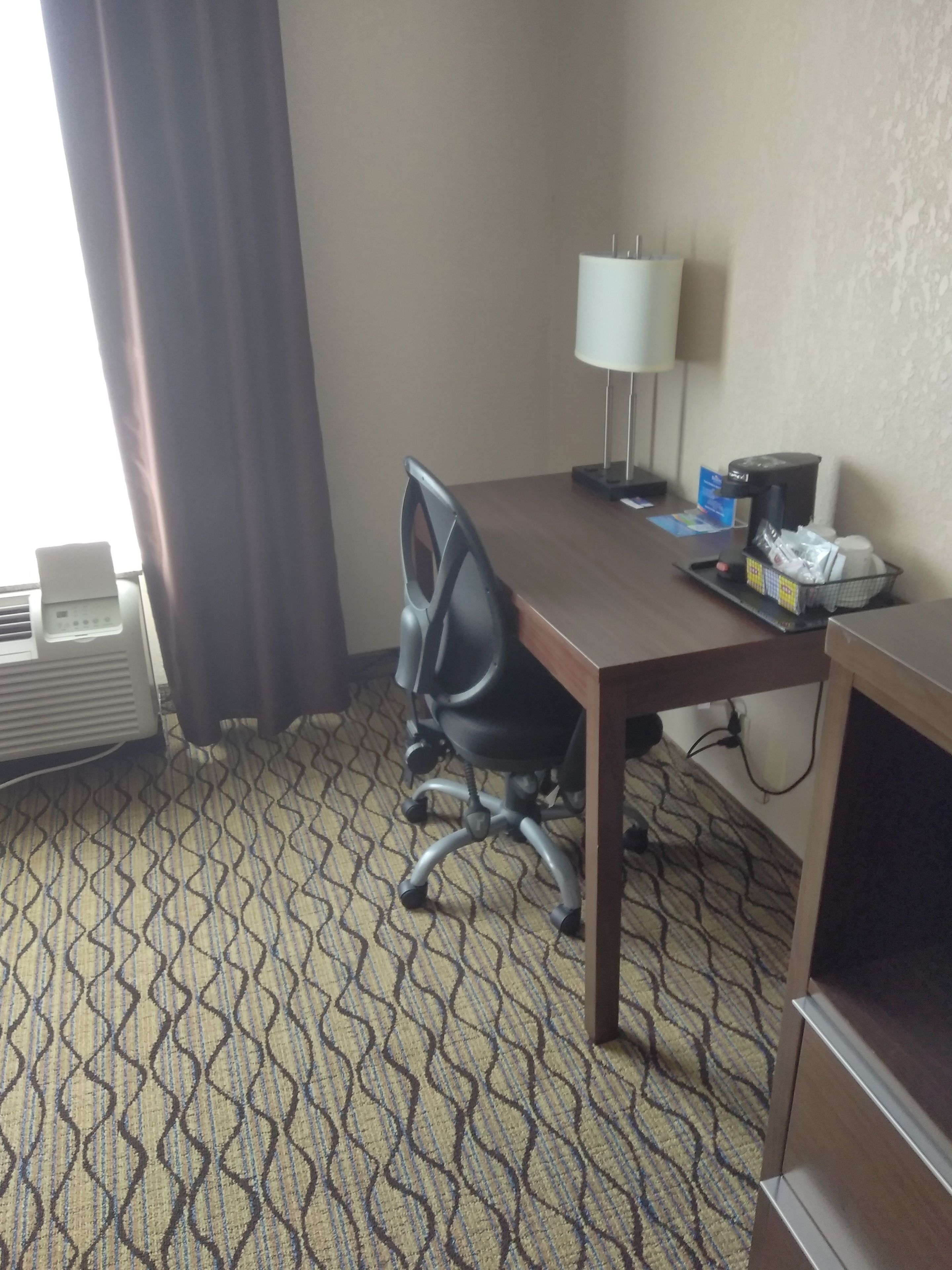 Une chambre d’hôtel équipée d’un bureau, d’une chaise, d’une lampe, d’un climatiseur et d’une moquette à motifs.