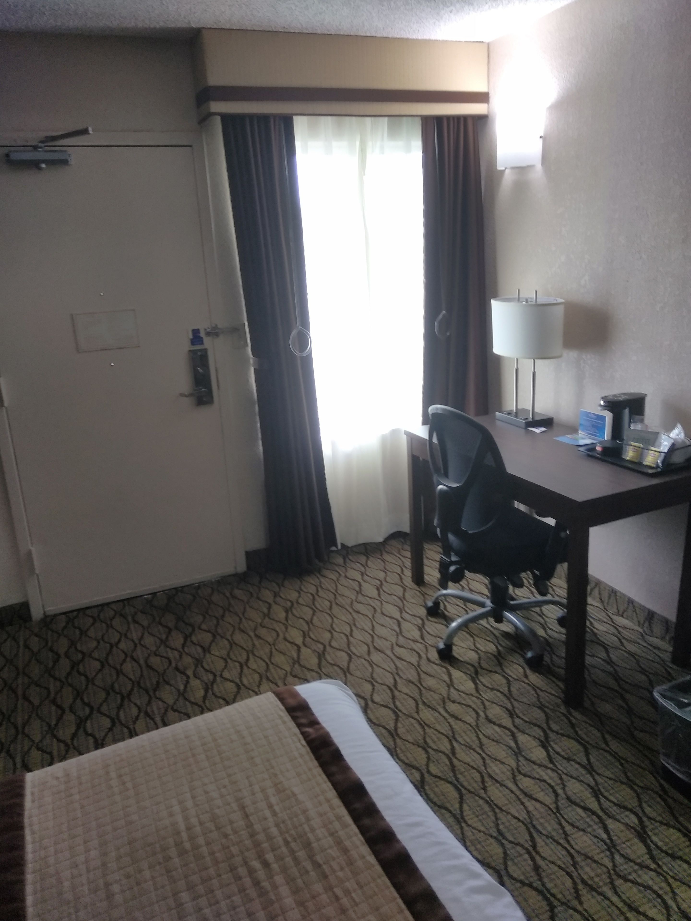 Habitación de hotel con cama, escritorio con silla, lámpara y ventana con cortinas.