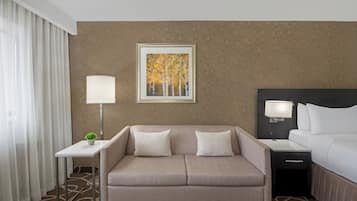 A hotel room with a sofa, a bed, a nightstand, a lamp, a framed picture on the wall, and a small table with a plant.