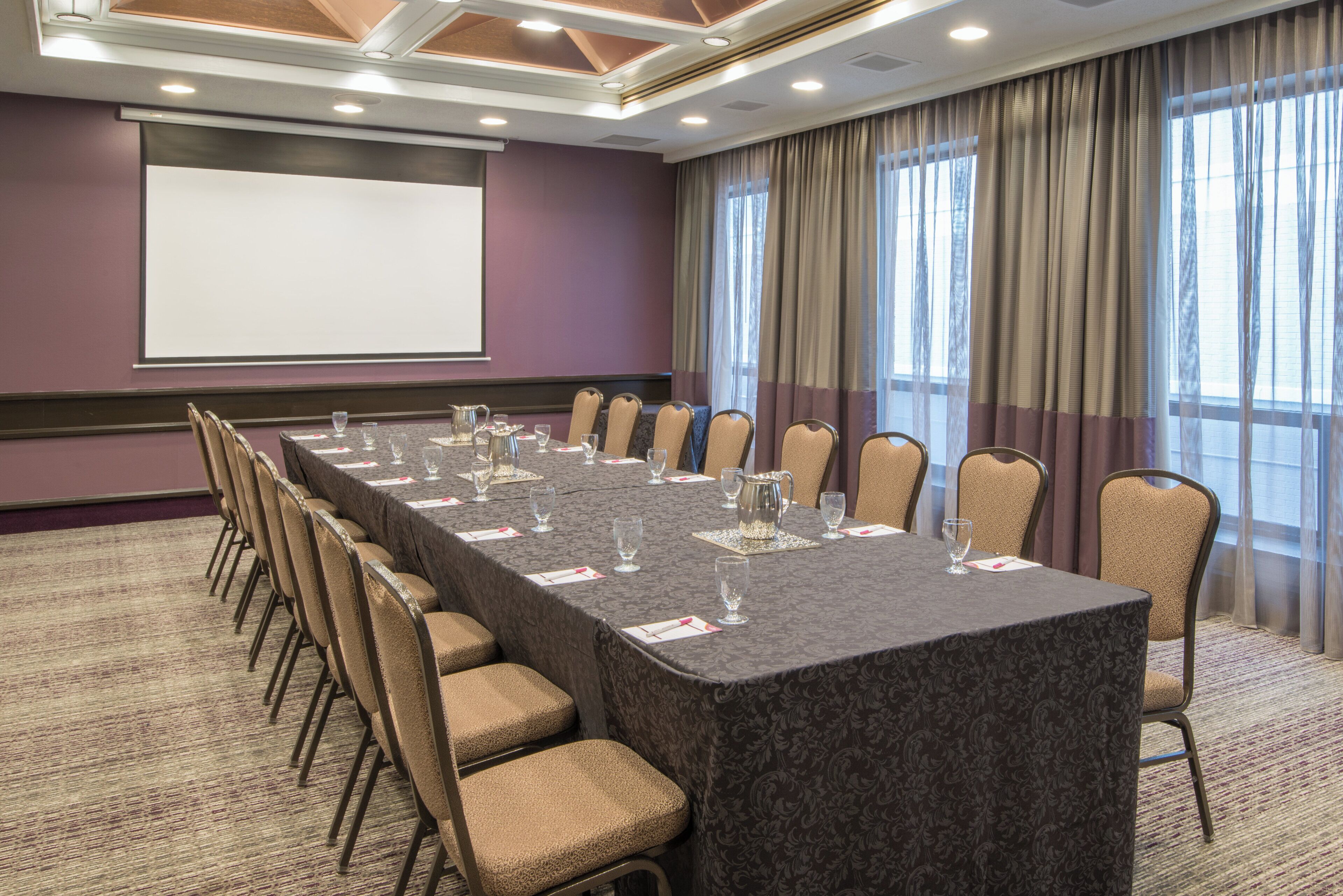 Una sala conferenze con un lungo tavolo, sedie, uno schermo per proiettore e ampie finestre con tende.