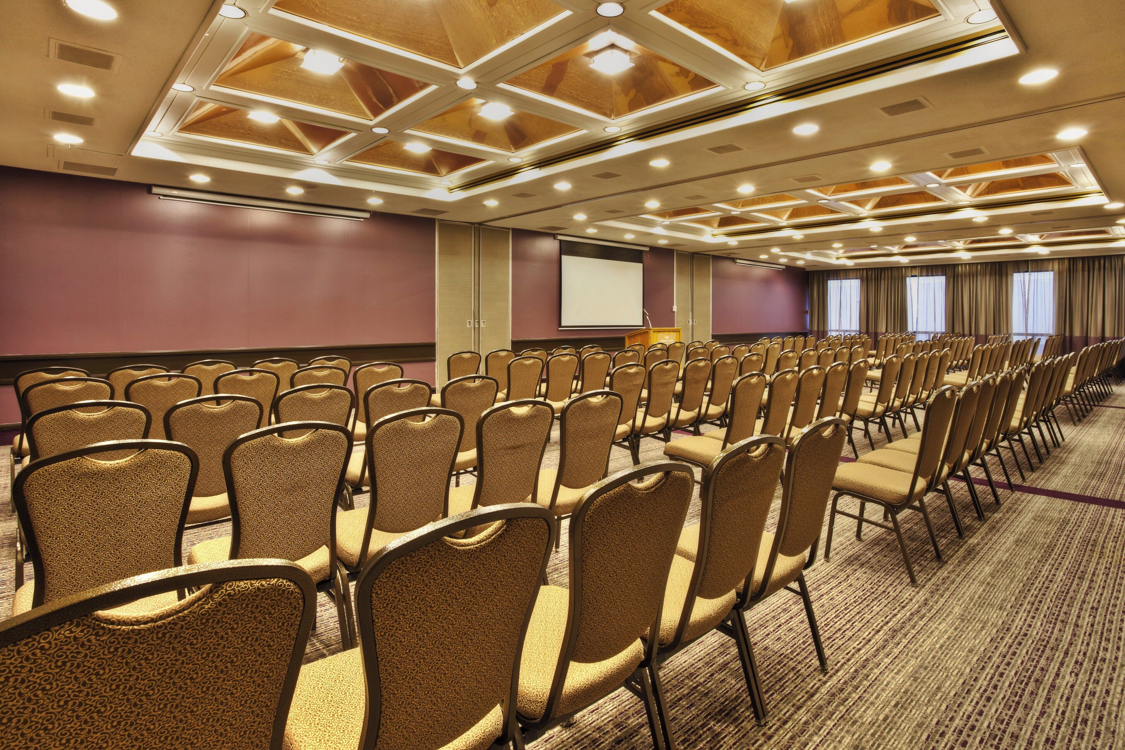 Un salón de conferencias con filas de sillas, una pantalla de proyector y un atril.