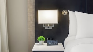 A neatly made bed with white linens, a digital clock displaying 12:05, a small potted plant, and a wall-mounted light fixture.