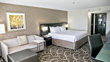 Quarto de hotel com cama, sofá, mesa pequena, cadeira e carpete estampado.
