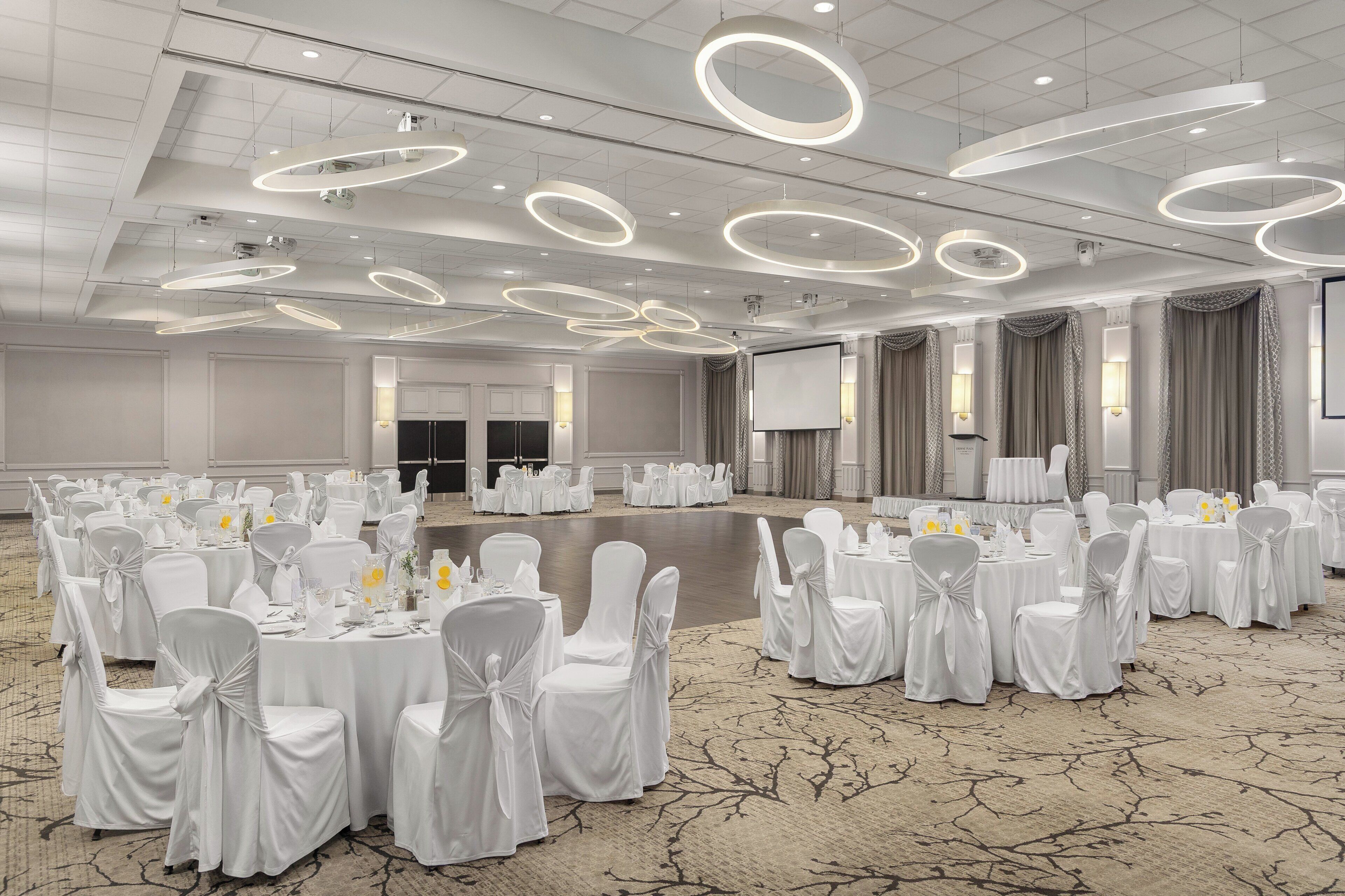 Un salón de banquetes con mesas redondas, sillas blancas y una pantalla grande.