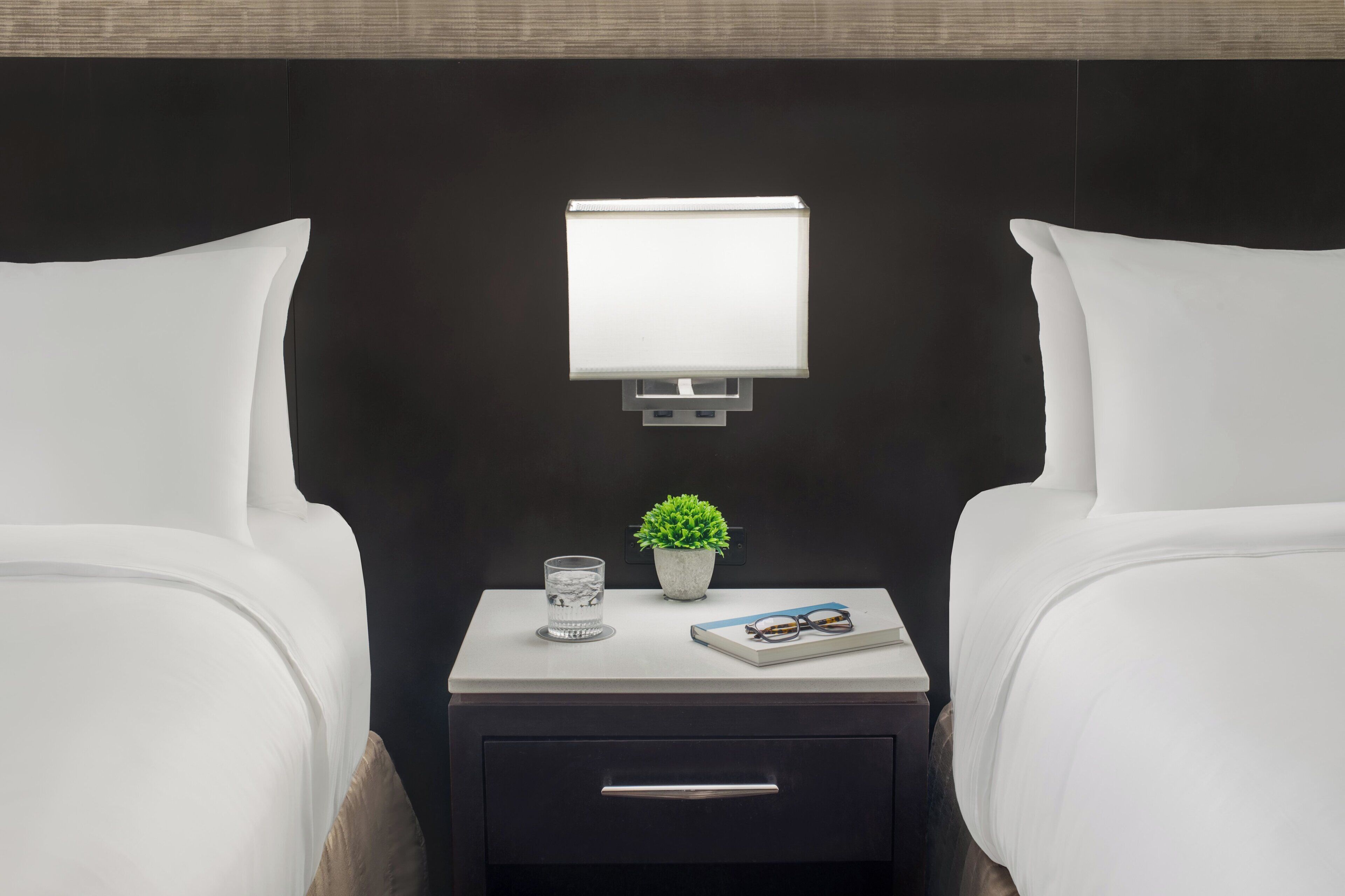 A hotel room with a bed, a bedside table with a lamp, a small potted plant, a glass of water, and a book.