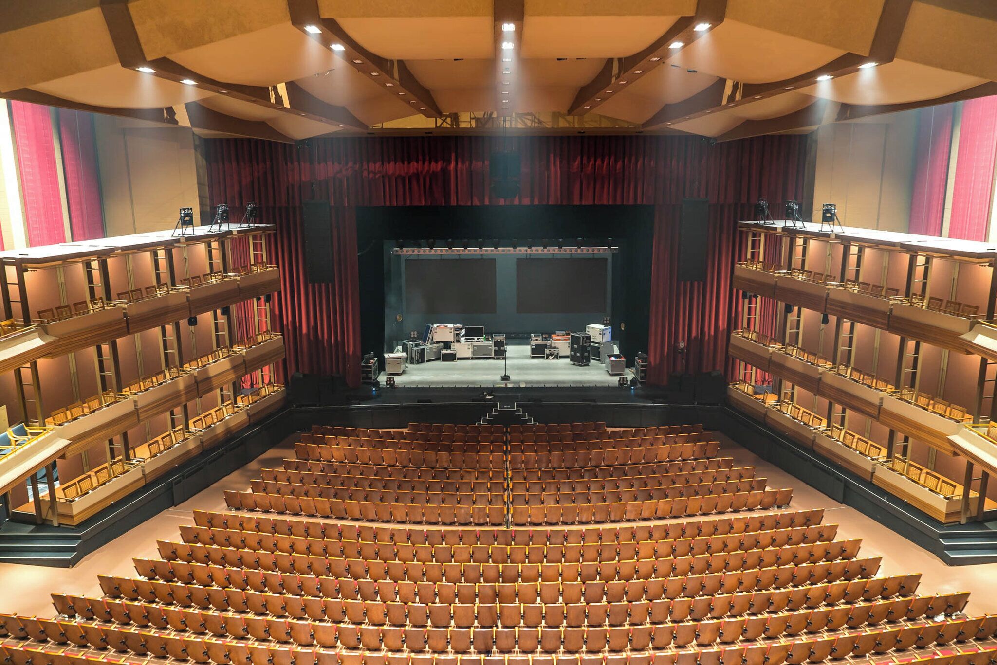 A large auditorium with tiered seating, a stage with lighting equipment, and a red curtain backdrop.
