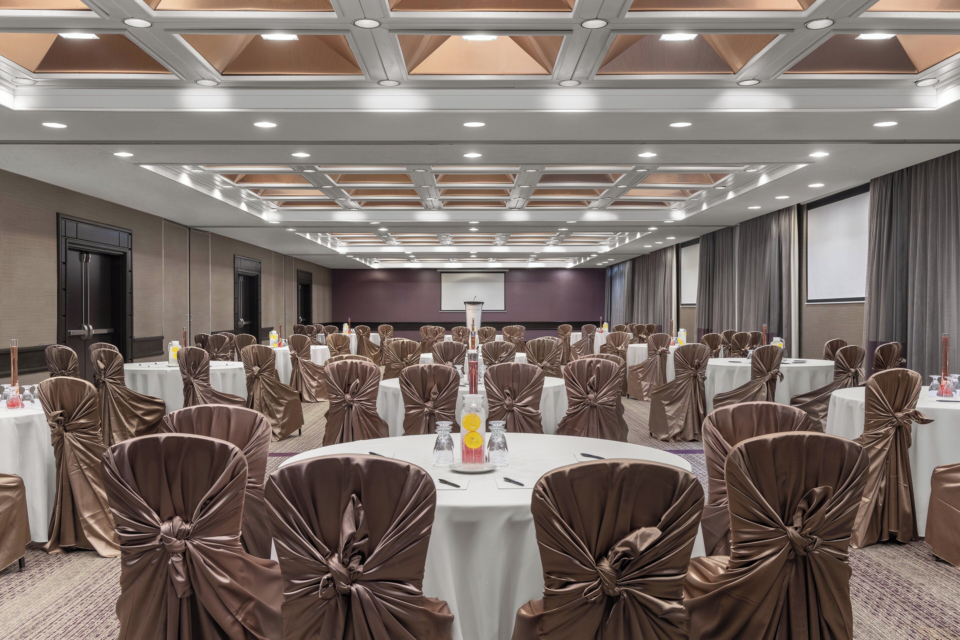 A banquet hall with round tables, chairs with brown sashes, and a ceiling with recessed lighting.