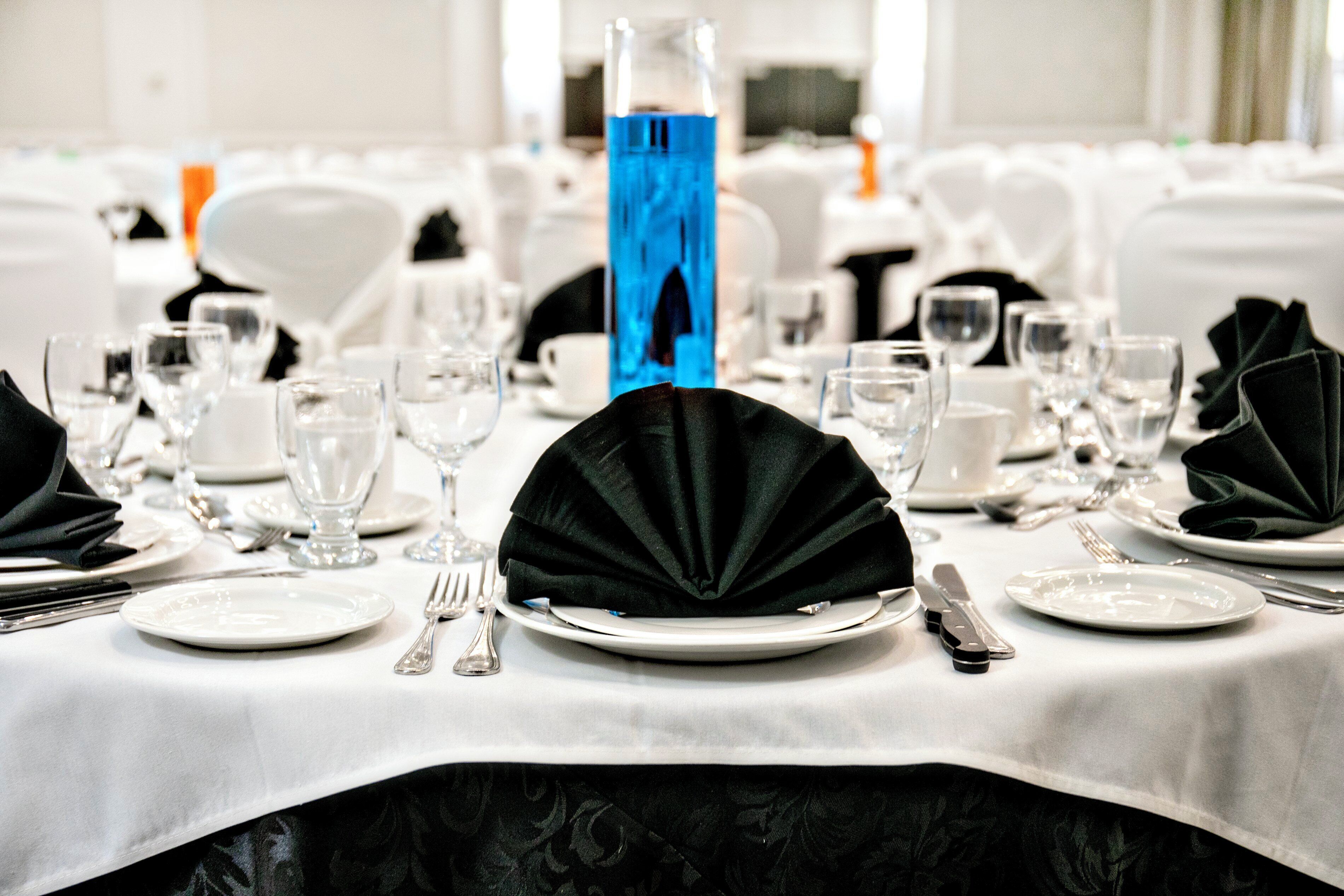 Mesa dispuesta para un evento formal con manteles blancos, servilletas negras y cristalería transparente.