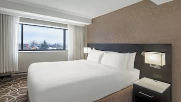 Quarto de hotel com uma cama grande, mesa de cabeceira com luminária, vista para edifícios e árvores pela janela, e um tapete estampado.