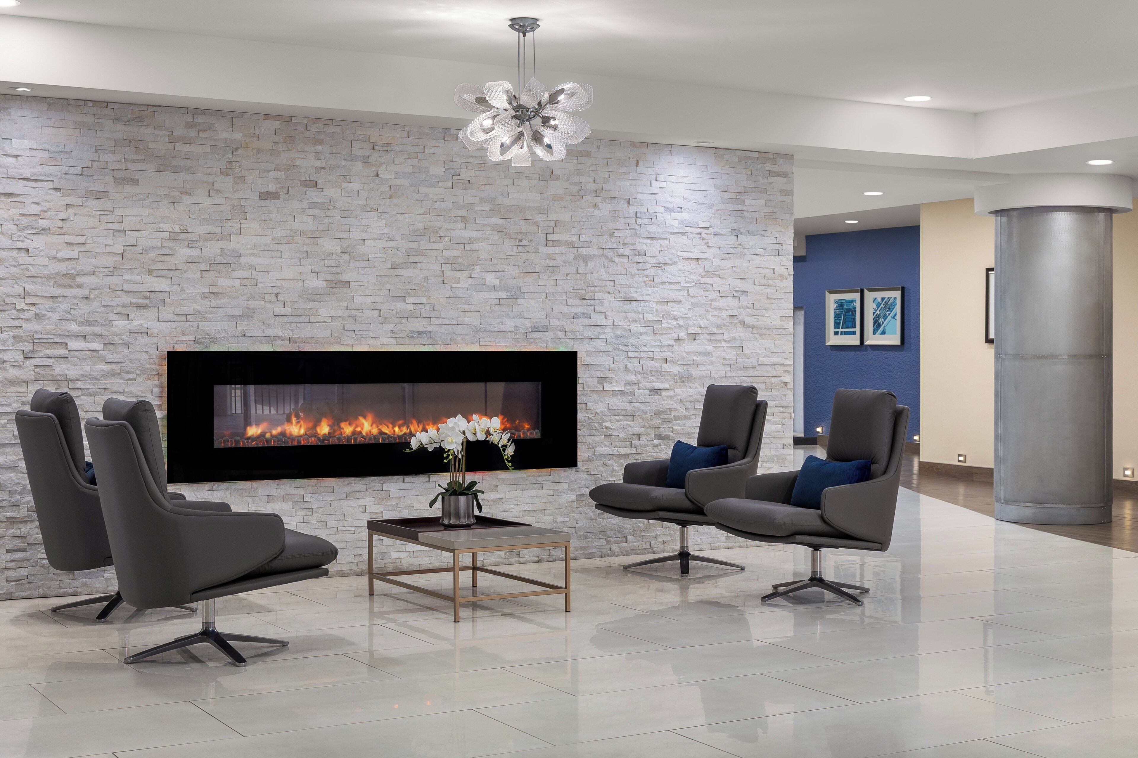 A modern lounge area with a stone wall, a flat-screen TV, and a fireplace. There are two grey lounge chairs and a small table with a vase of flowers.