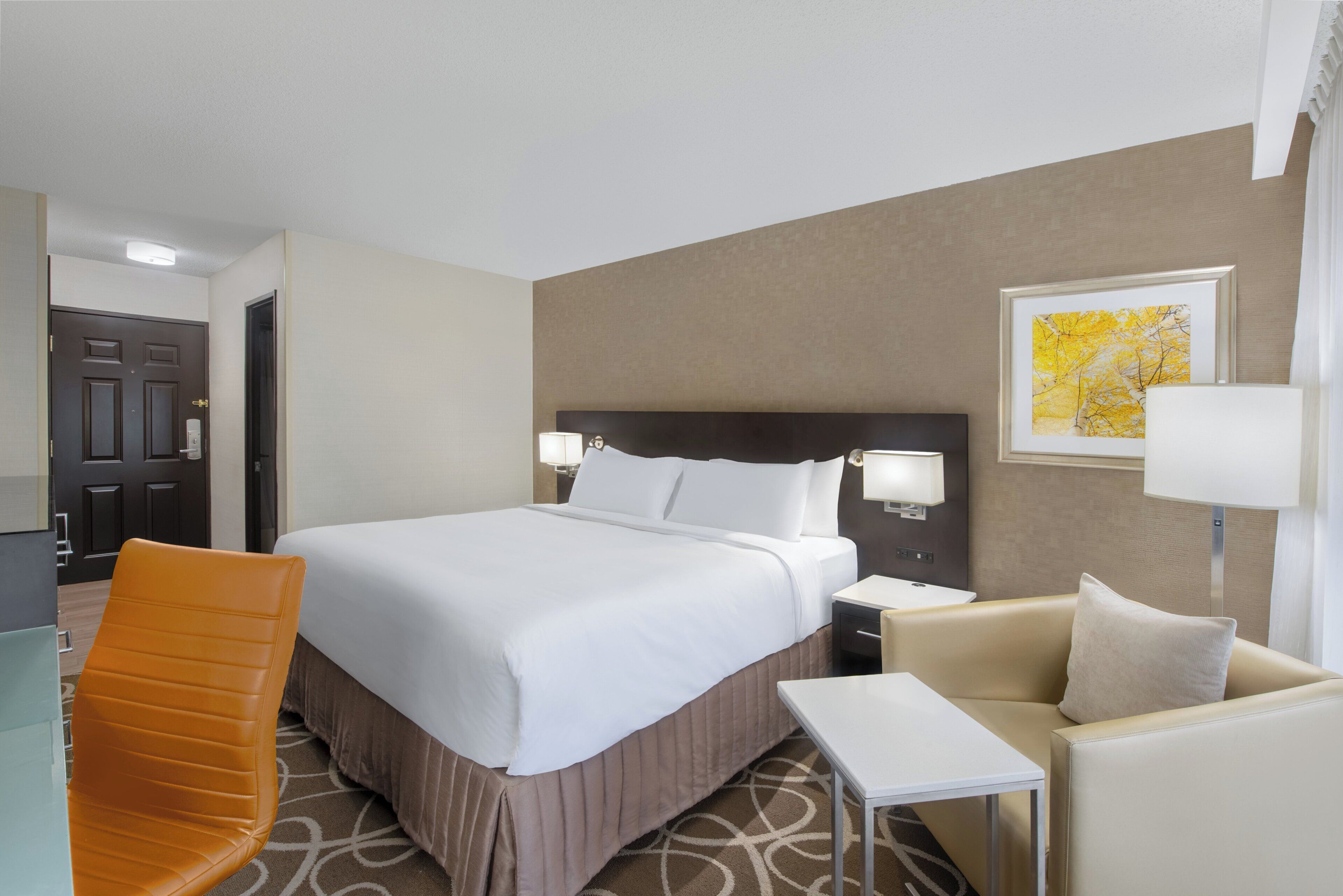 A hotel room with a bed, a chair, a small table, and a nightstand with a lamp.