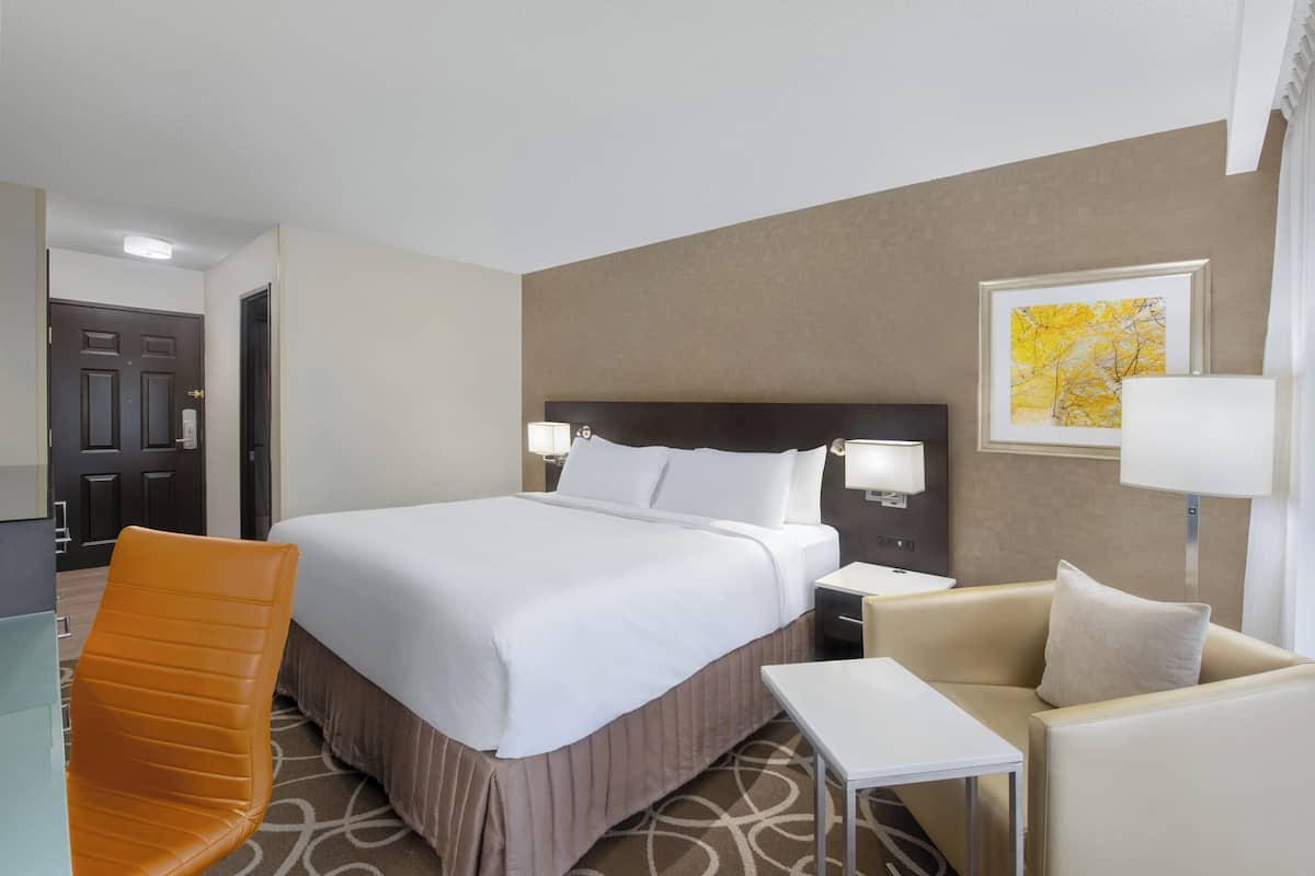 A hotel room with a bed, a chair, a small table, and a nightstand with a lamp.
