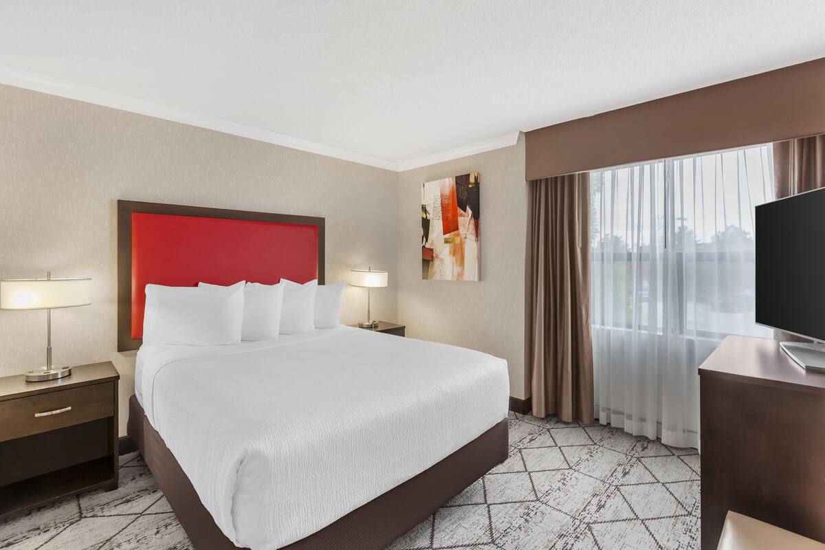 A hotel room with a large bed, a red headboard, a flat-screen TV, a desk, and a window with curtains.